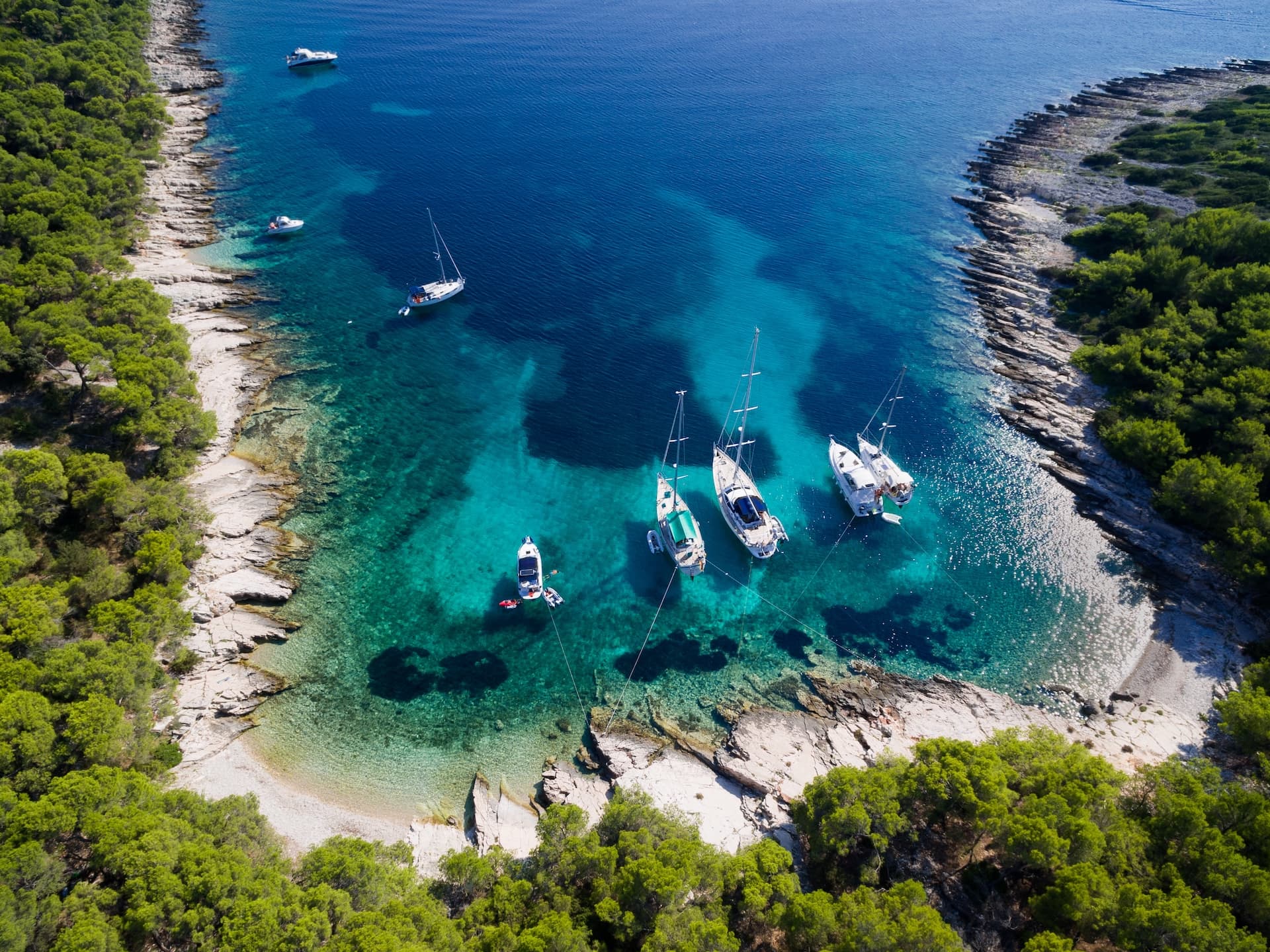 Aerial view of sailboats and motorboats anchored in turquoise cove near forested, rocky coastline of Otok Žirje.