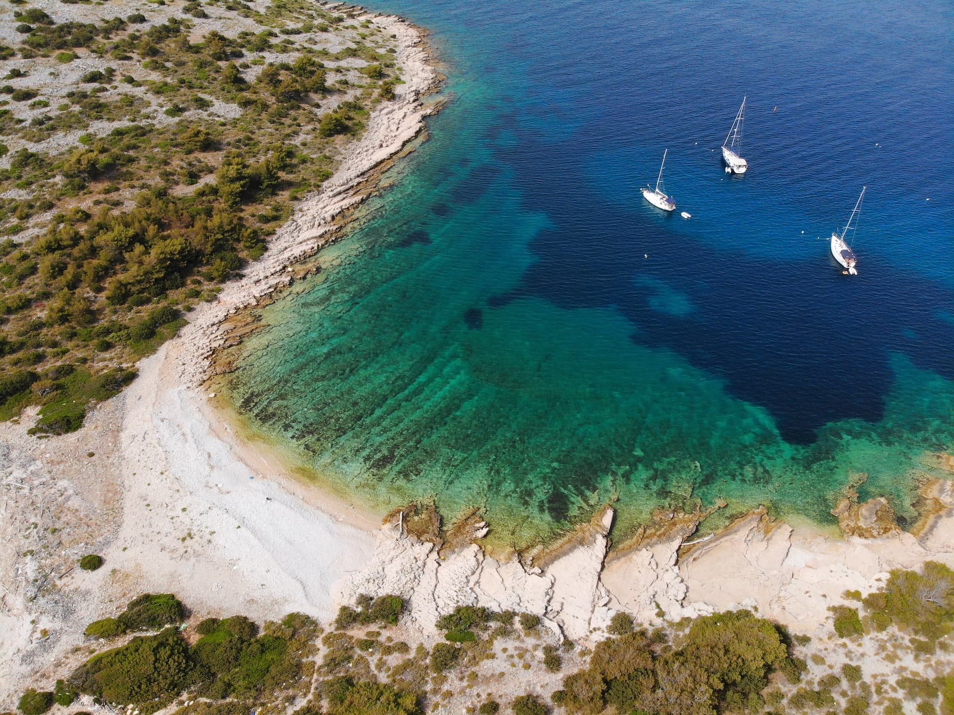 Three sailboats anchored near a rocky, scrub-covered island shore with turquoise and deep blue water.