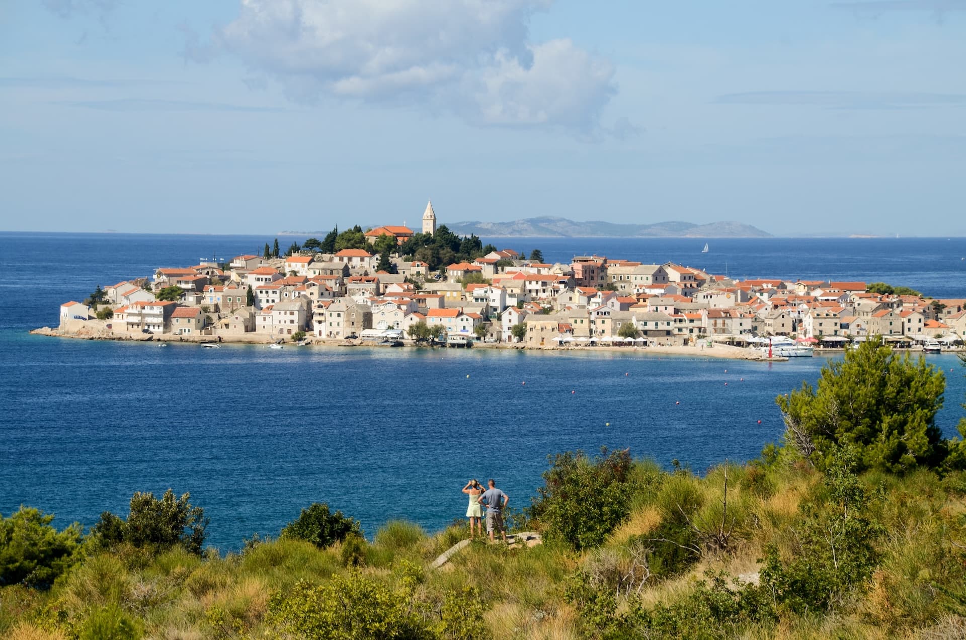Coastal town with terracotta roofs on a peninsula across blue water, viewed from a grassy hill.
