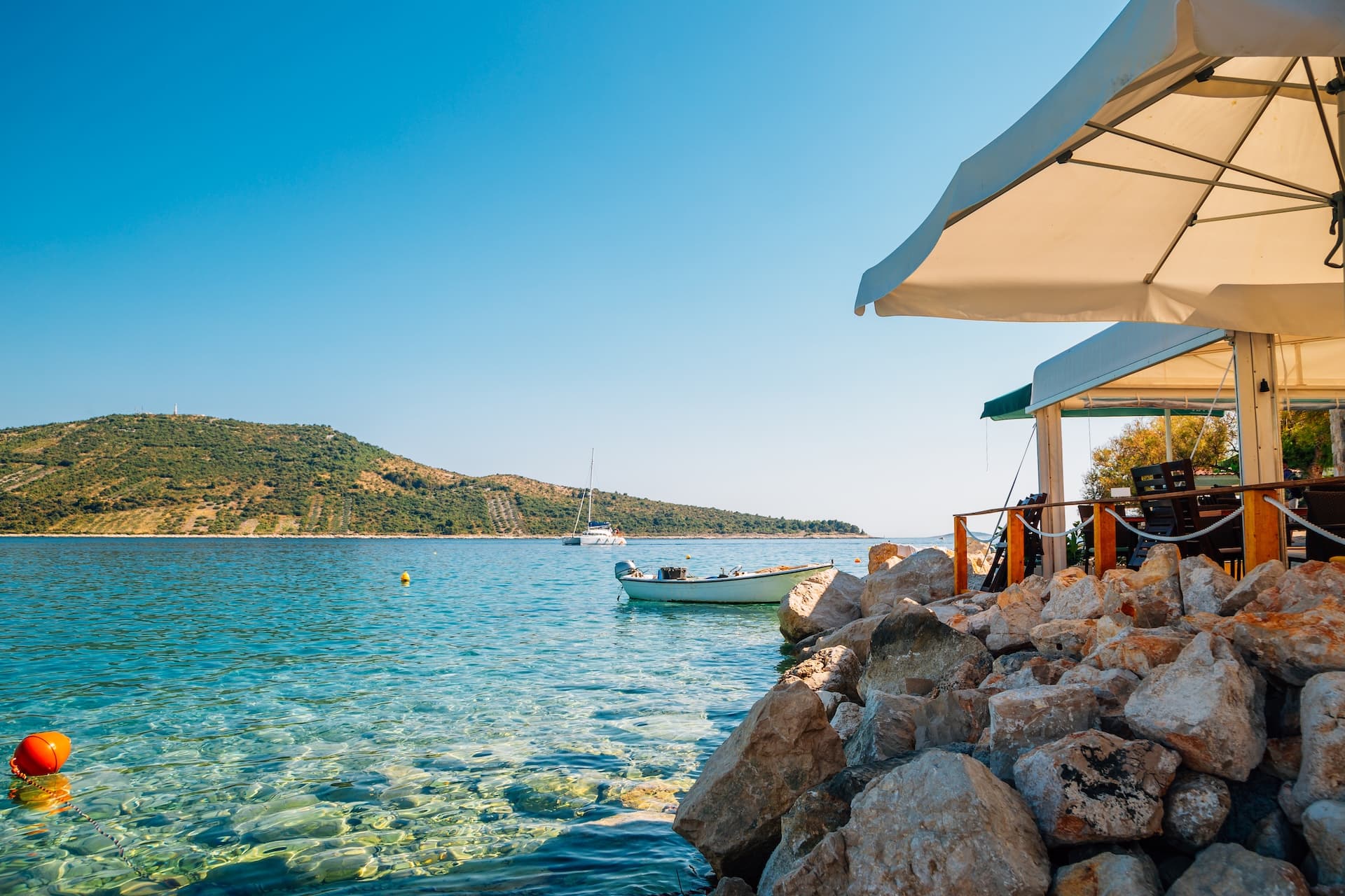 Outdoor restaurant seating by rocky shore with clear water, boat, and green hill in Primošten.