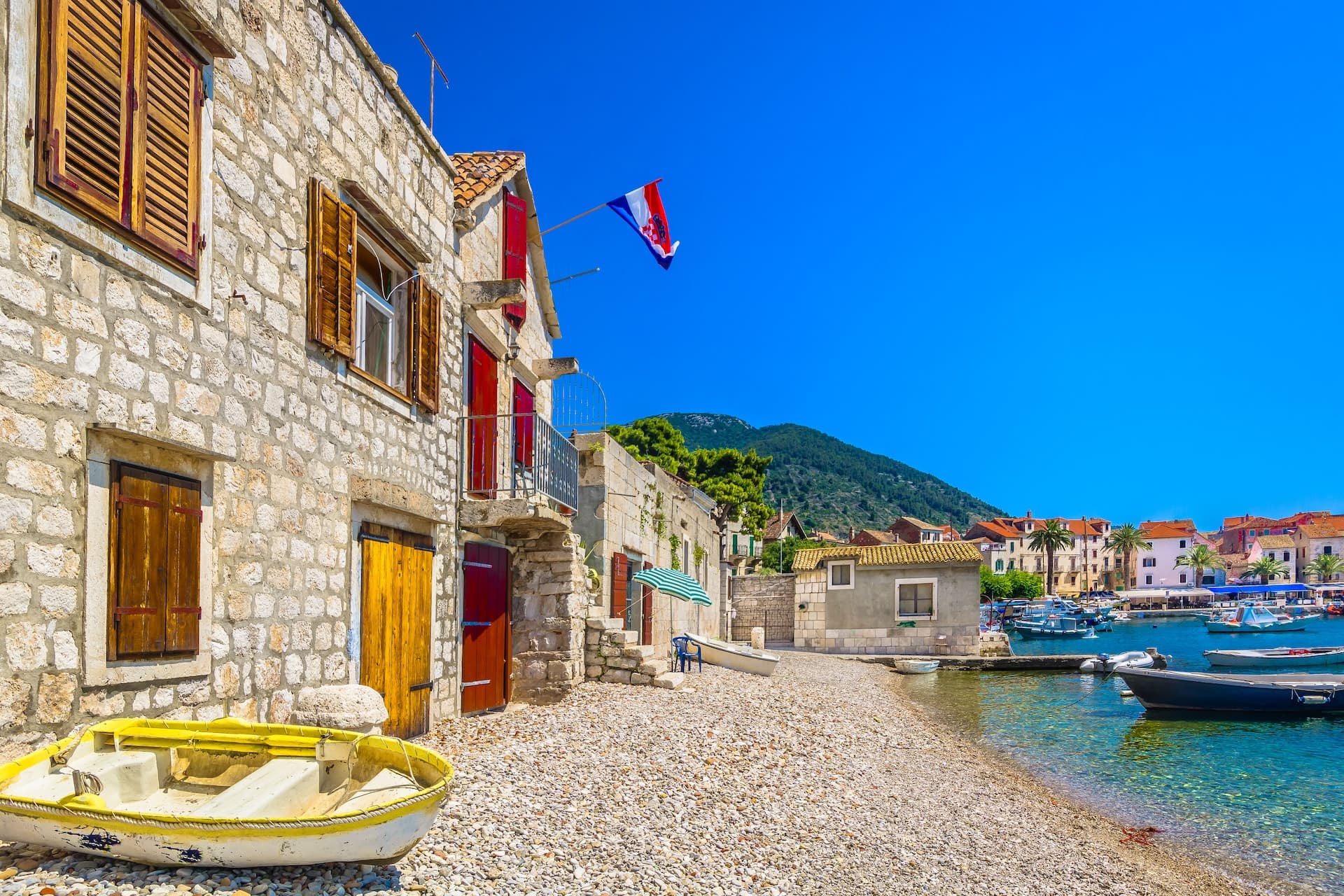 Stone houses on pebble beach with boats in clear water near Komiza Island.
