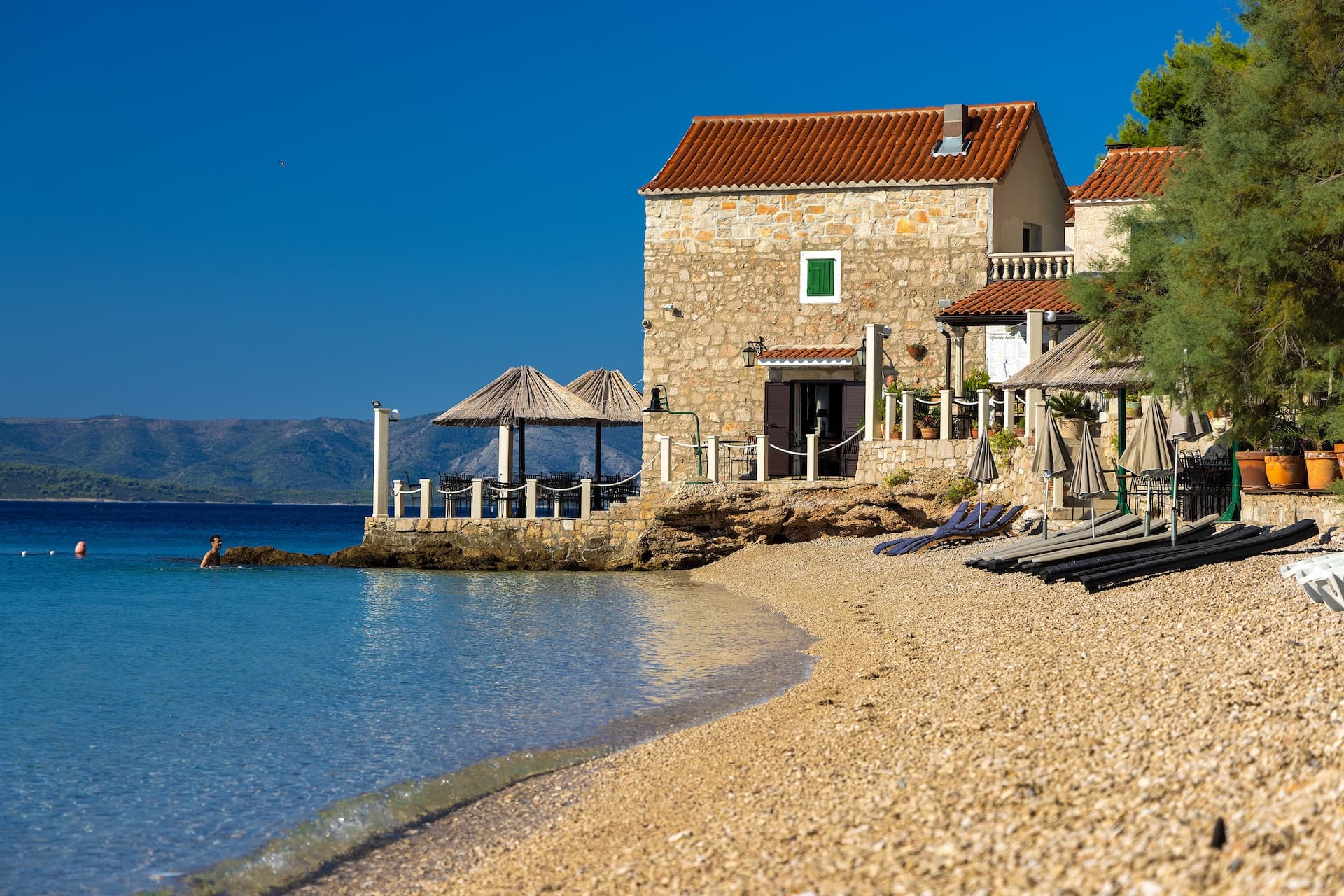 Stone building by pebble beach with clear blue water and distant green mountains.