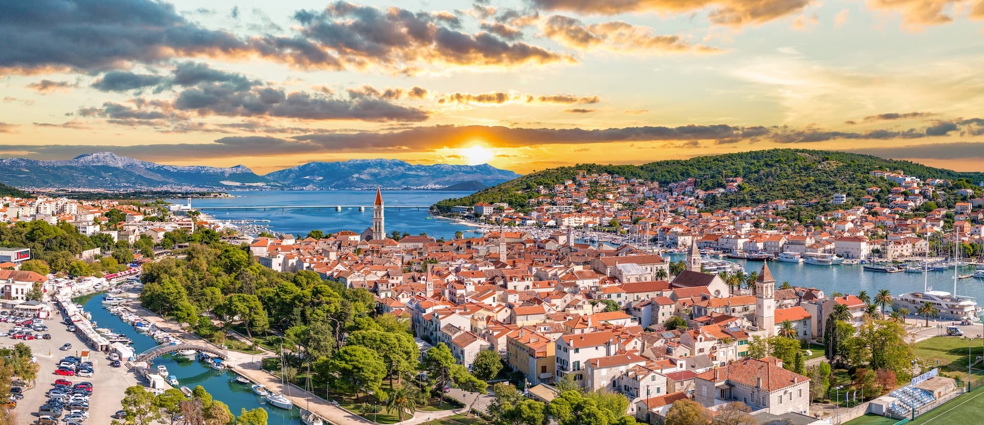 Coastal town with red roofs and a bell tower at sunset, overlooking mountains and the sea.