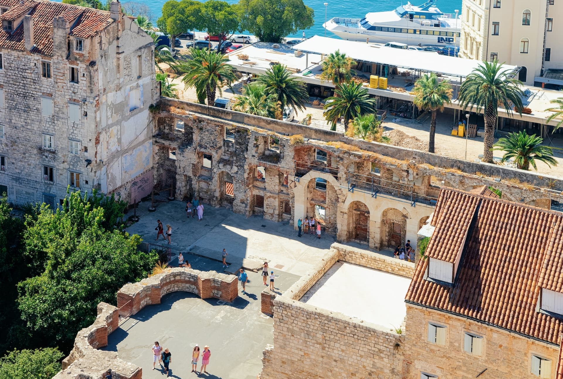 Historic stone ruins courtyard with palm trees, overlooking a white yacht on blue coastal water.