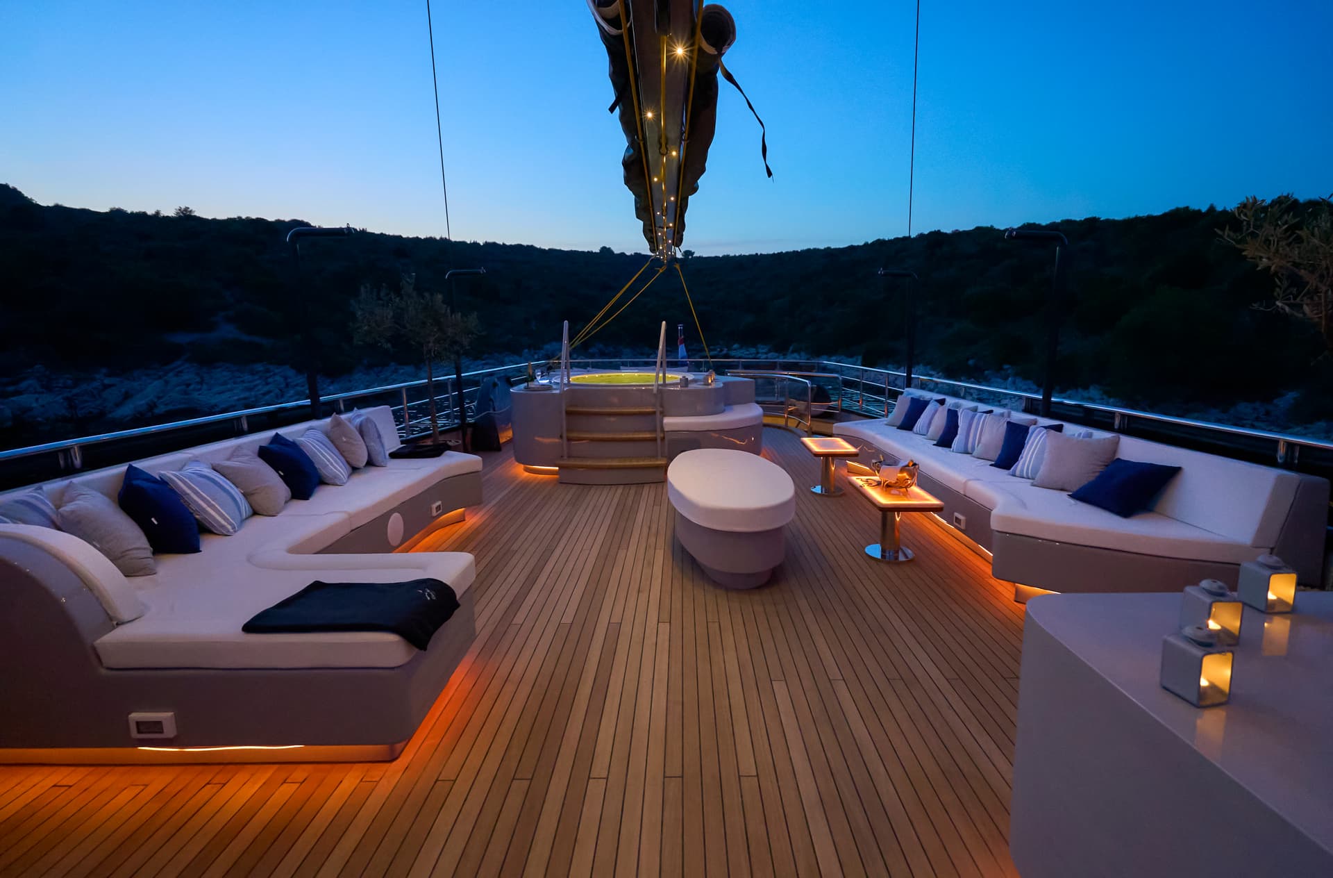 Luxury yacht deck seating area with hot tub at dusk overlooking dark coastline.