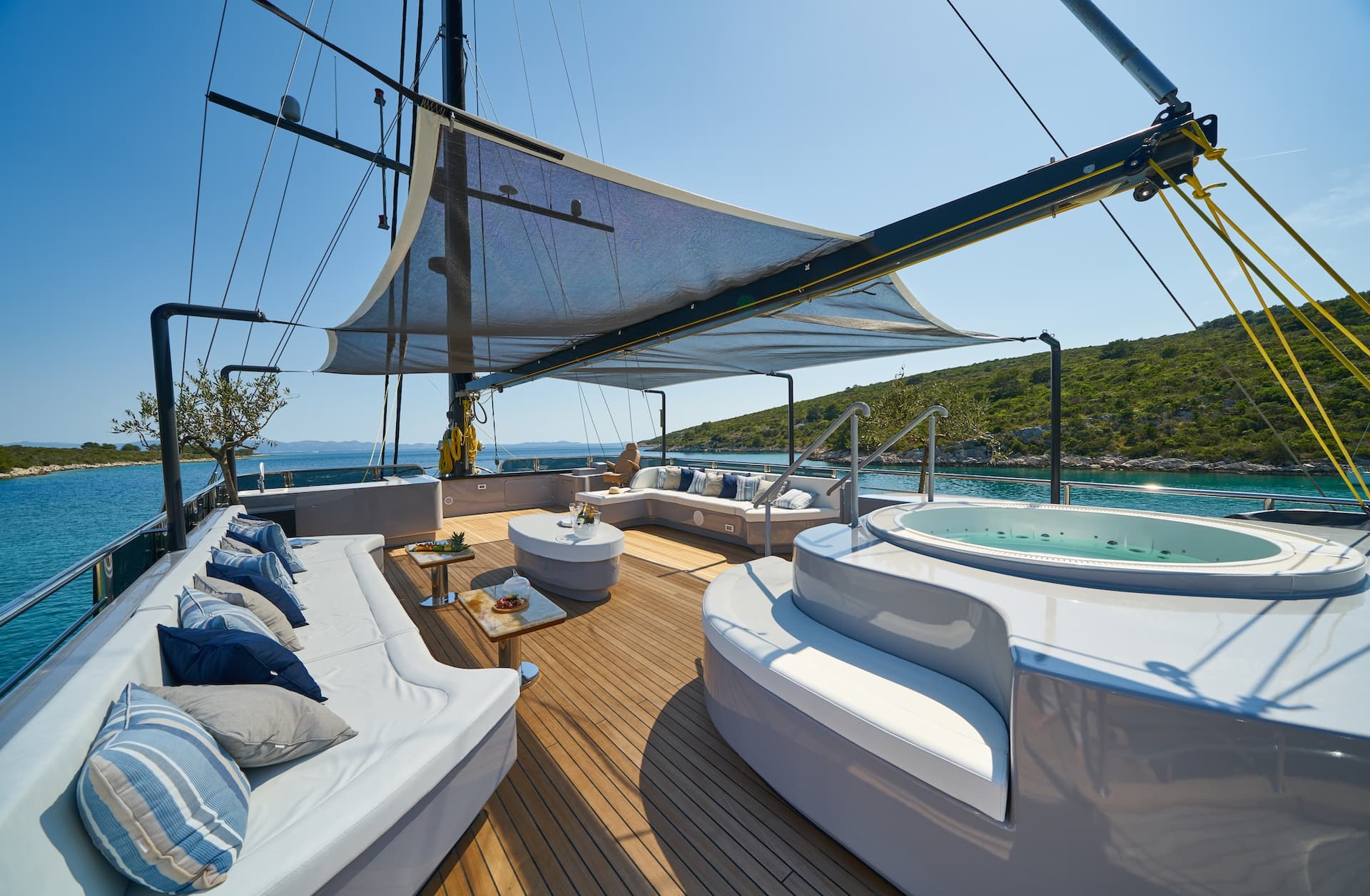 Yacht deck with jacuzzi, white seating, and sunshade anchored near green coastline.