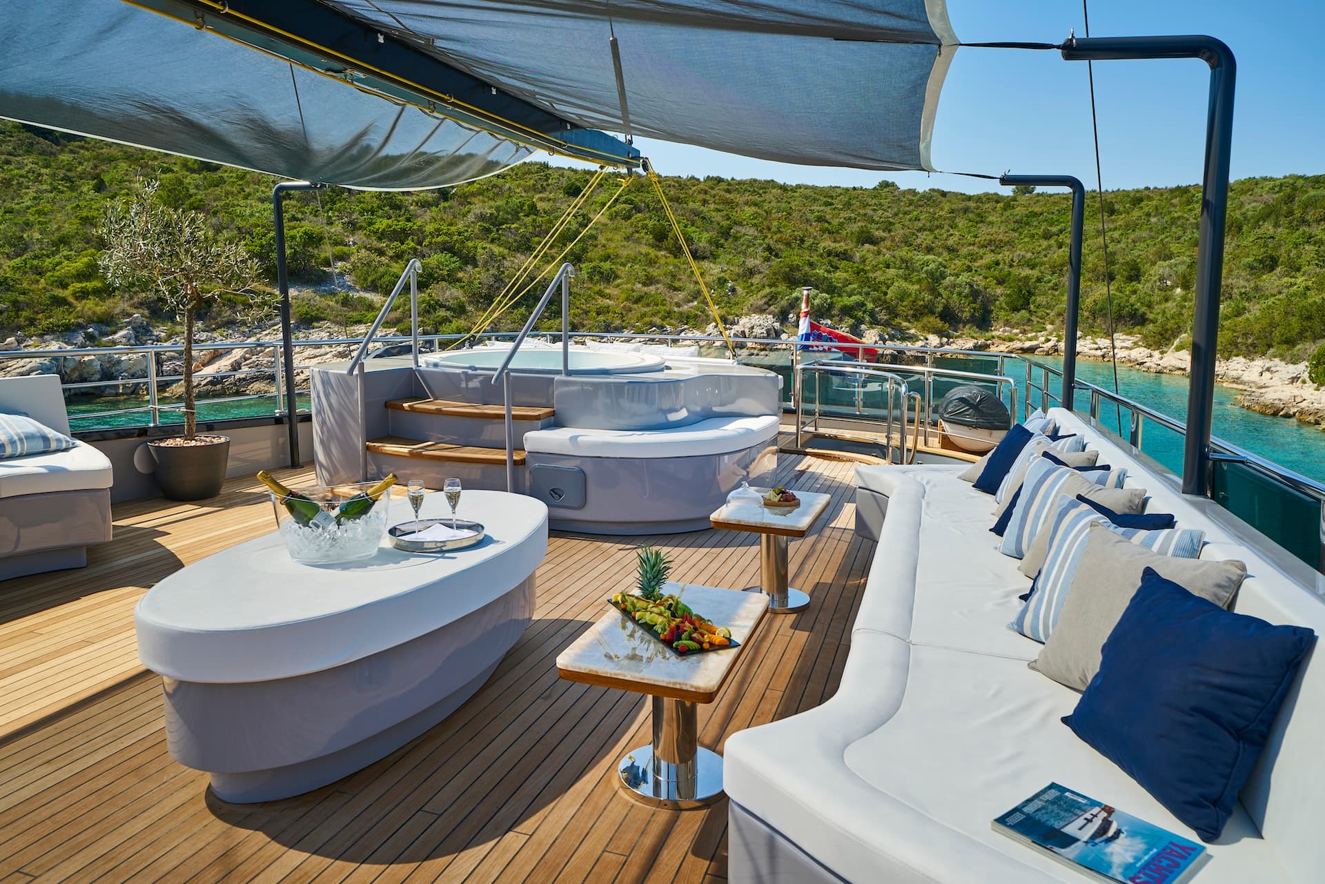 Yacht deck with hot tub, champagne, and fruit platter anchored near lush green coastline.