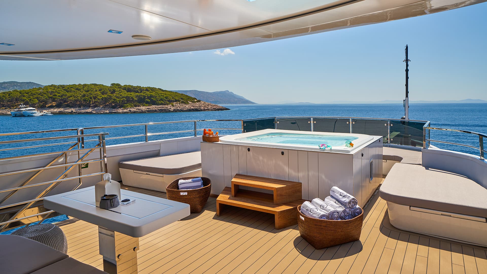 Jacuzzi deck on a yacht with sun loungers overlooking a wooded island and blue sea.