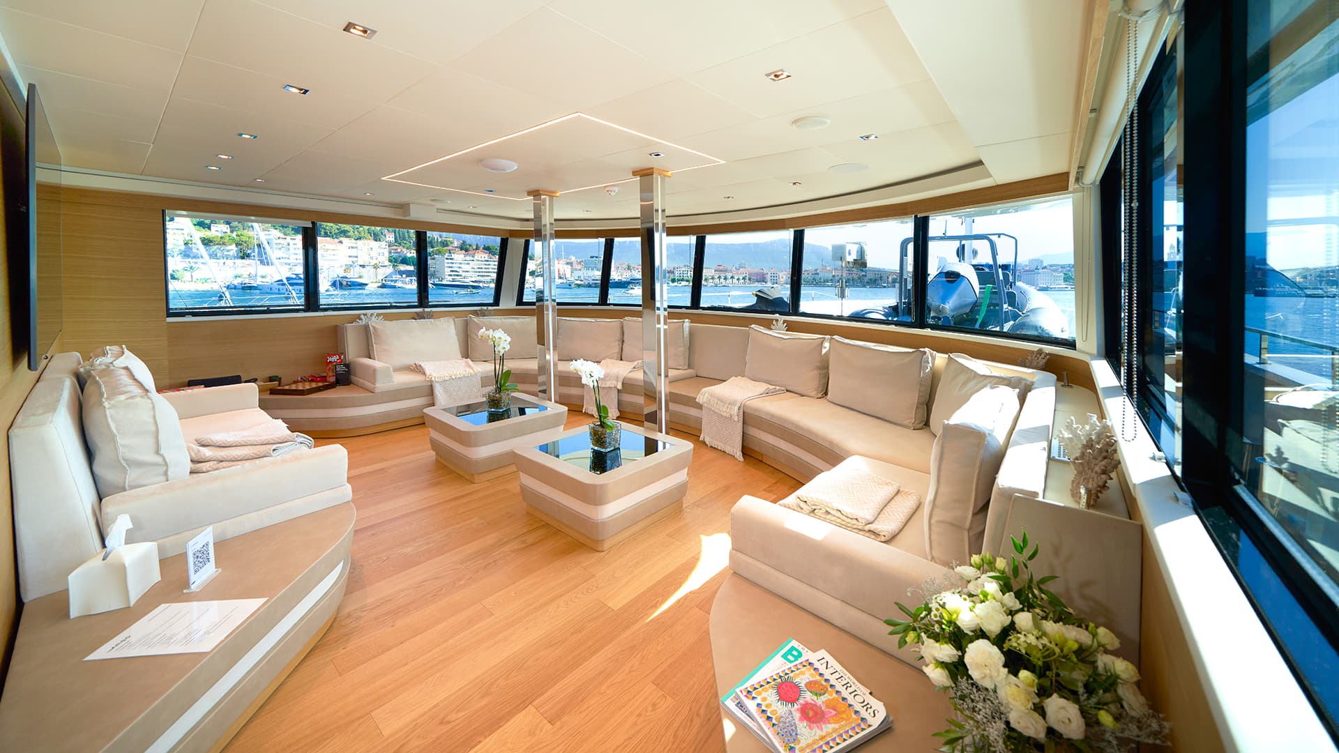 Yacht lounge with beige seating, wood floors, and view of a coastal town harbor.
