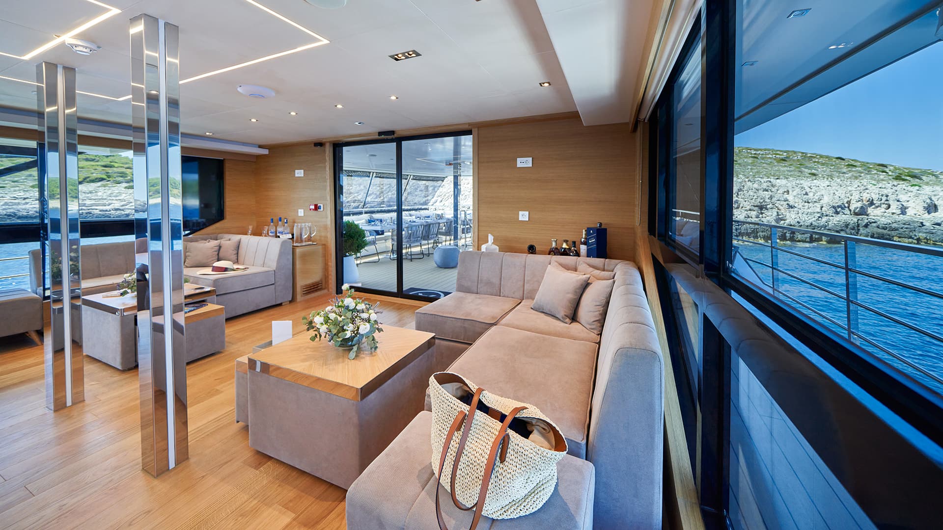 Modern yacht interior lounge with wood paneling, seating, and view of rocky coastline and blue water.