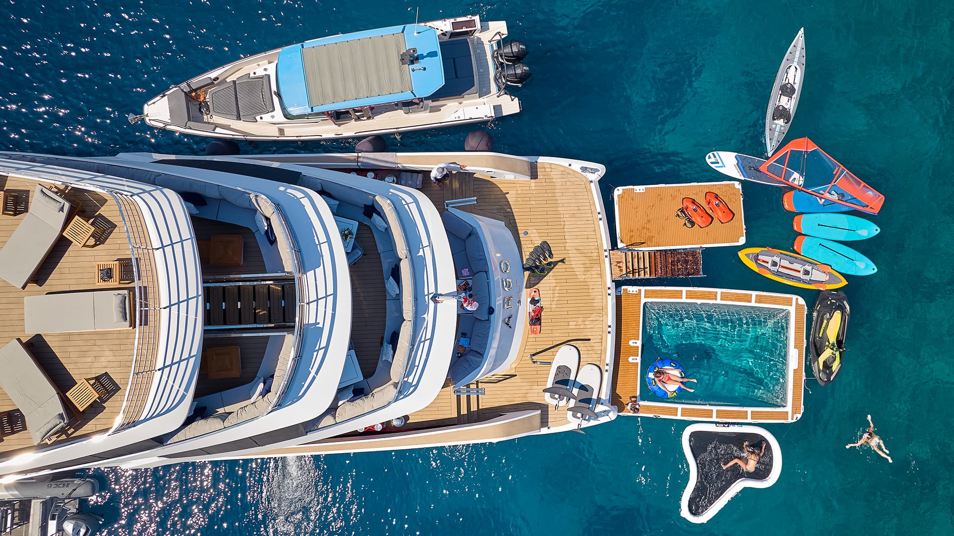 Aerial view of a large yacht deck with sun loungers, water toys, and a floating pool in clear blue sea.