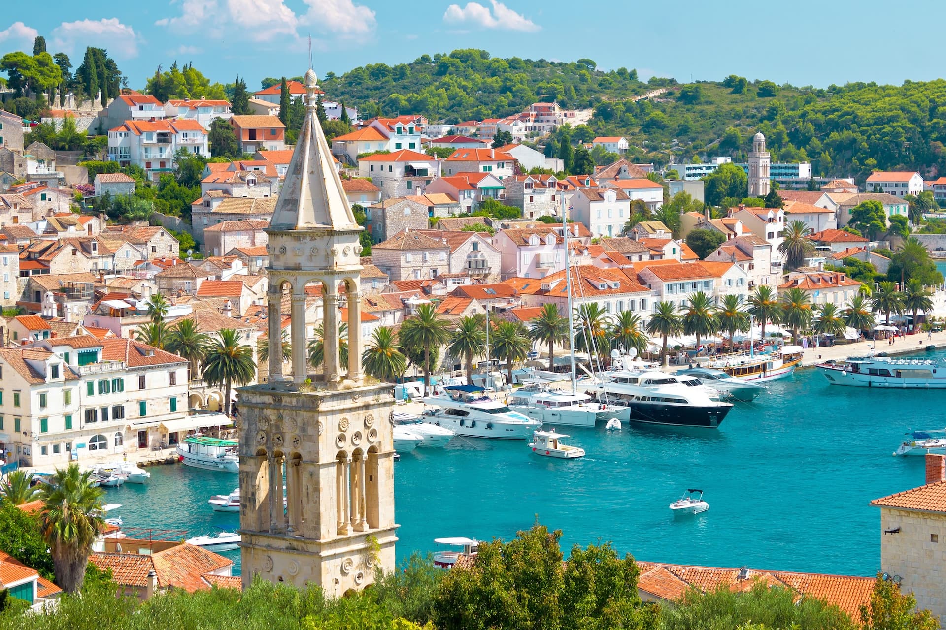 Stone bell tower overlooking Hvar harbor with white buildings, palm trees, and yachts on turquoise water.