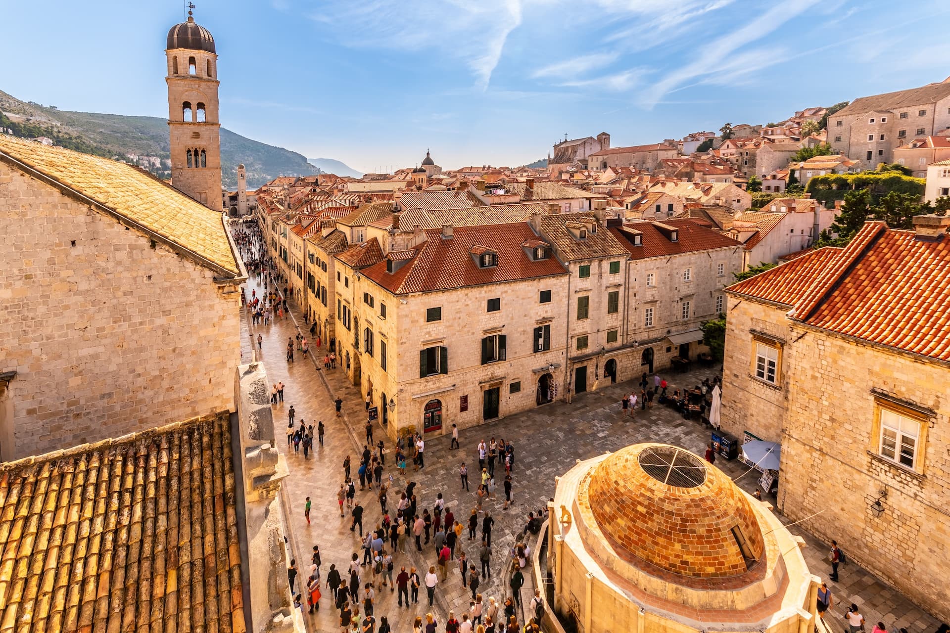 Crowded historic street and square in Dubrovnik with stone buildings and bell tower.