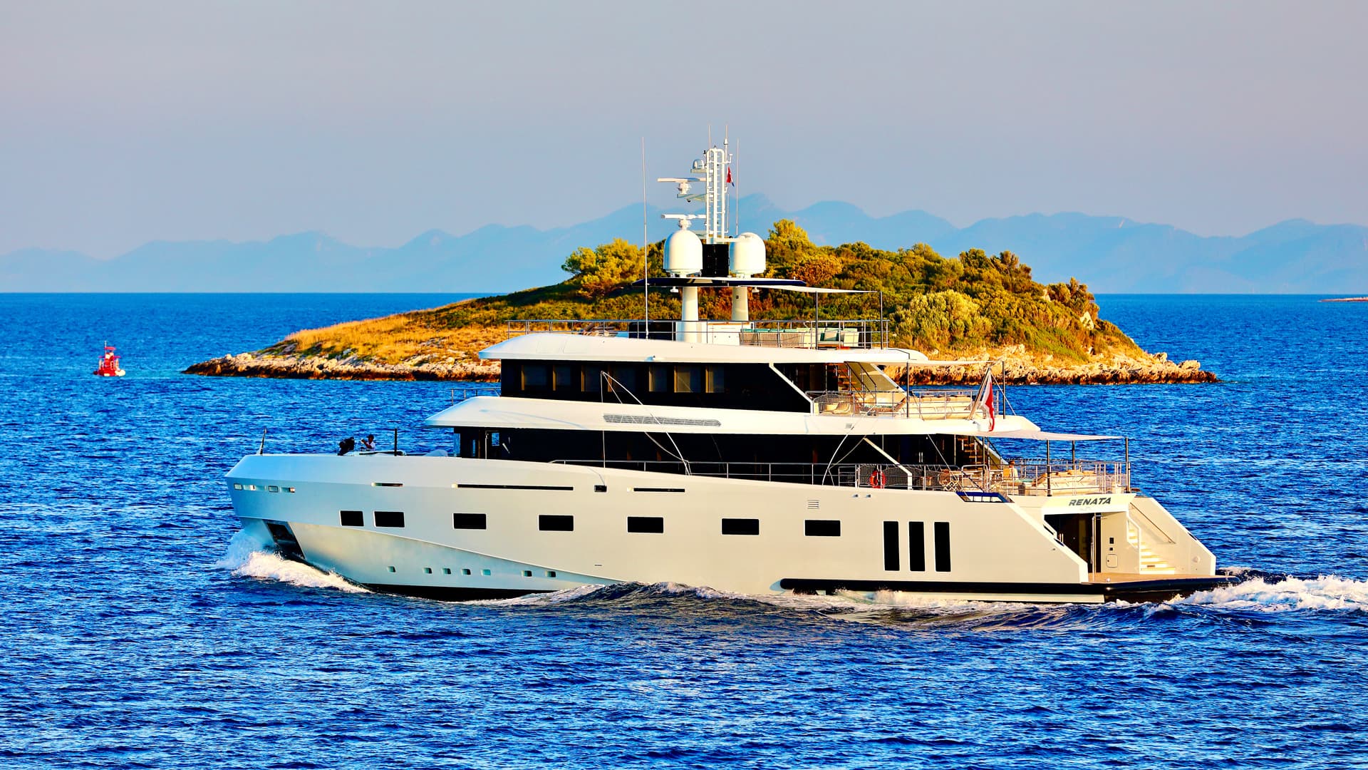 White motor yacht named RENATA cruising on blue sea near a small, scrub-covered island.