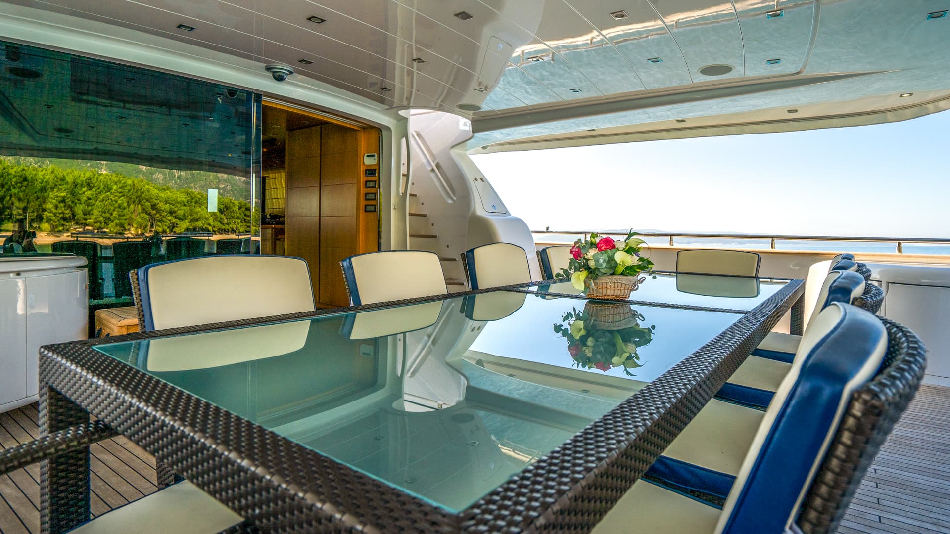 Outdoor dining area on a yacht with wicker chairs, glass table, and flowers, overlooking trees and the sea.