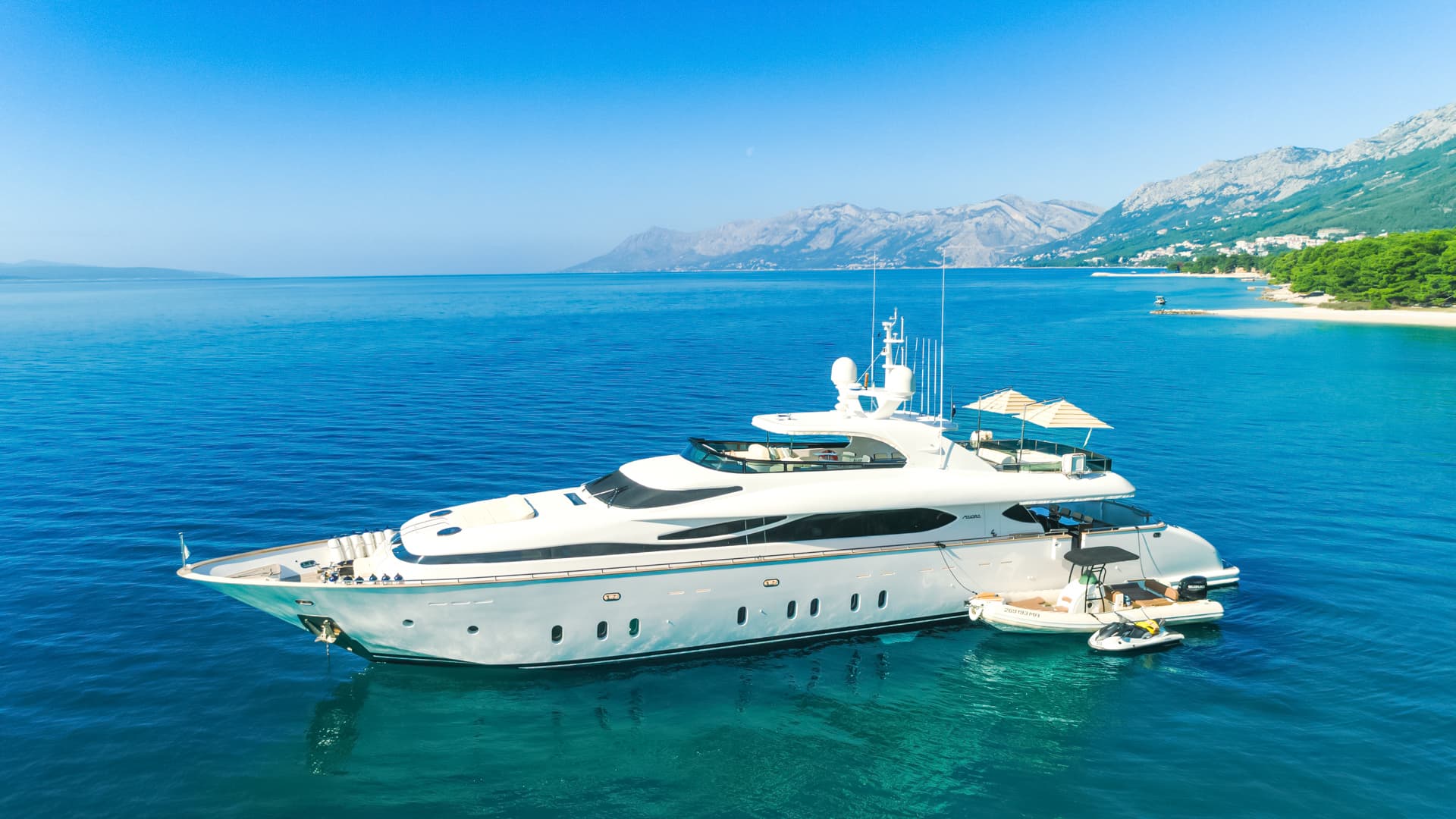 Luxury yacht anchored in clear blue sea near a mountainous coastline with a small beach.