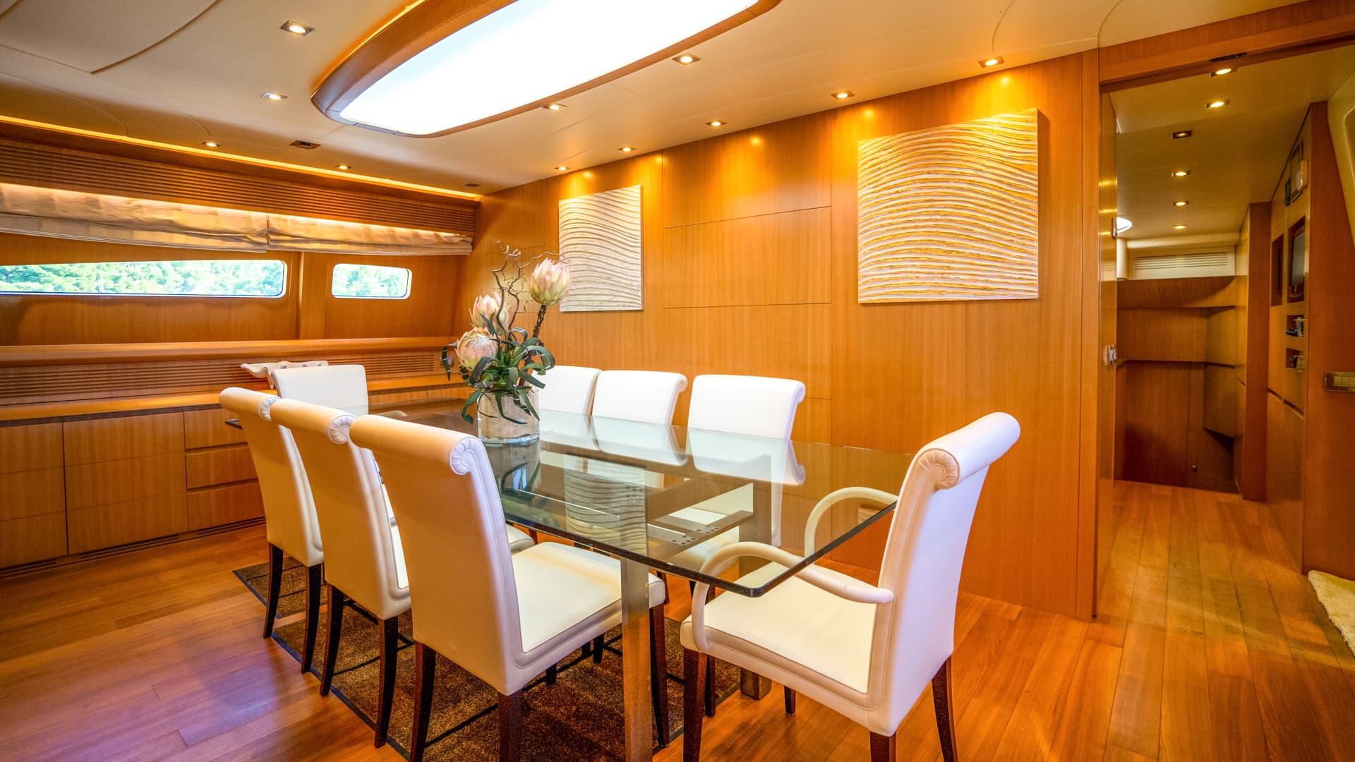 Dining area with glass table, white chairs, and wood paneling inside a yacht or boat.