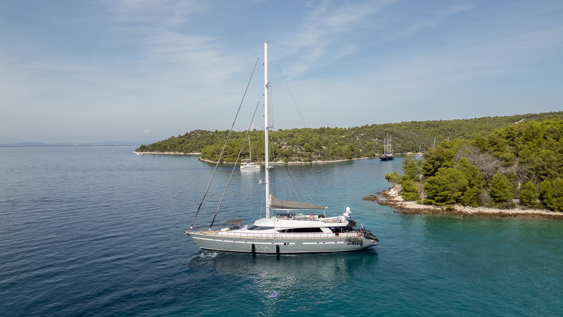 Large sailboat navigating clear blue water near a heavily forested coastline.