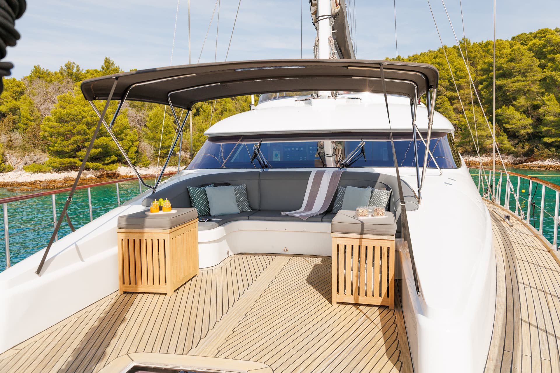 Bow lounge area of a white yacht with teak deck, anchored near a lush green coastline.