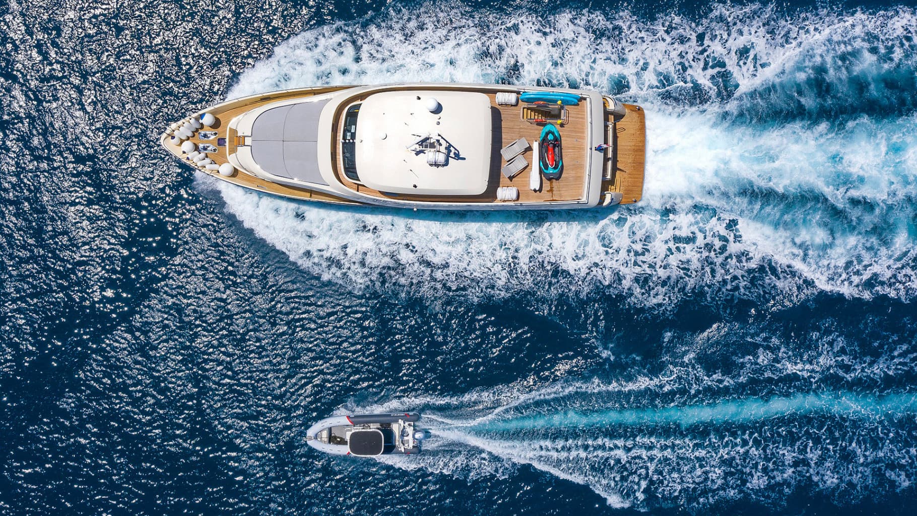 Yacht and small tender boat speeding across deep blue, sunlit ocean water, aerial view.