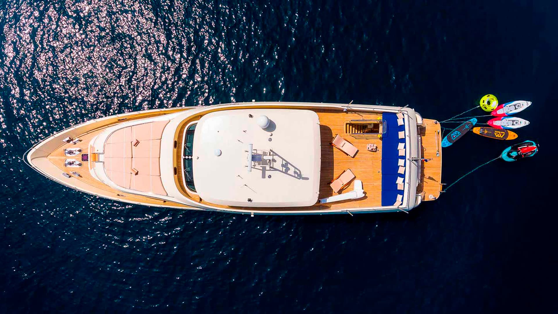 Yacht anchored on dark blue water with water toys floating off the stern, aerial view.