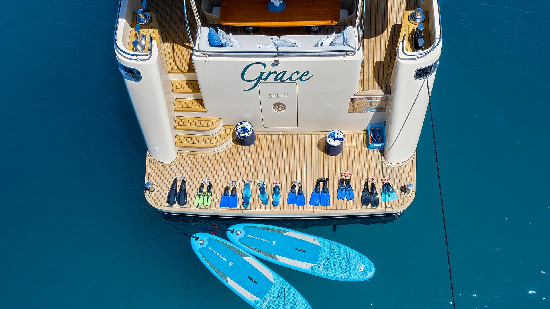 Yacht stern with "Grace SPLIT" lettering, swim fins, and paddleboards on blue water