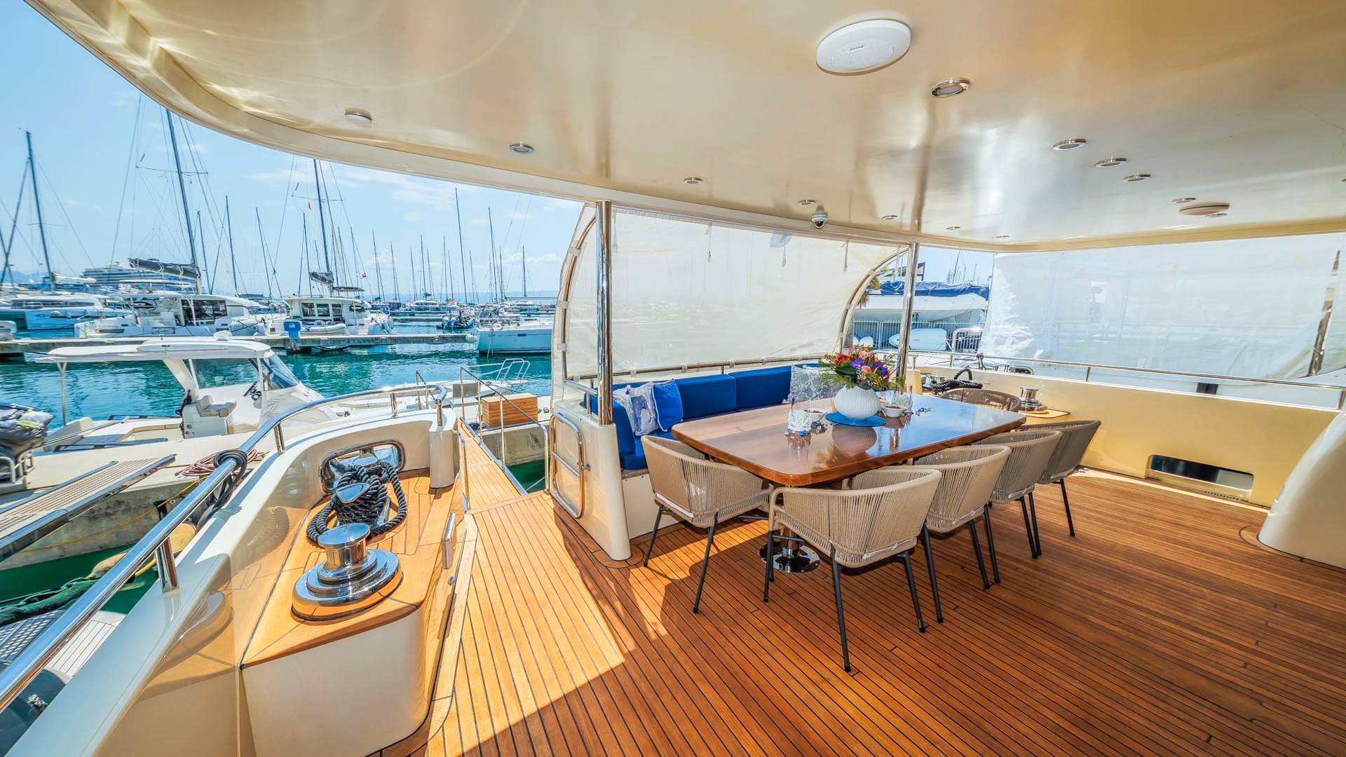 Yacht aft deck dining area with teak flooring overlooking a sunny marina full of sailboats.