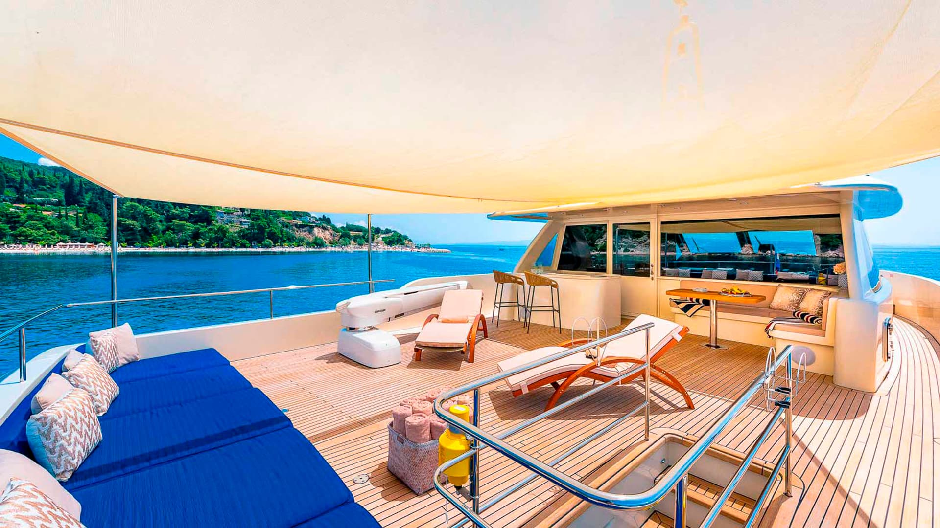 Flybridge deck of a yacht with teak flooring, blue cushions, and sunshade overlooking a green, hilly coastline.