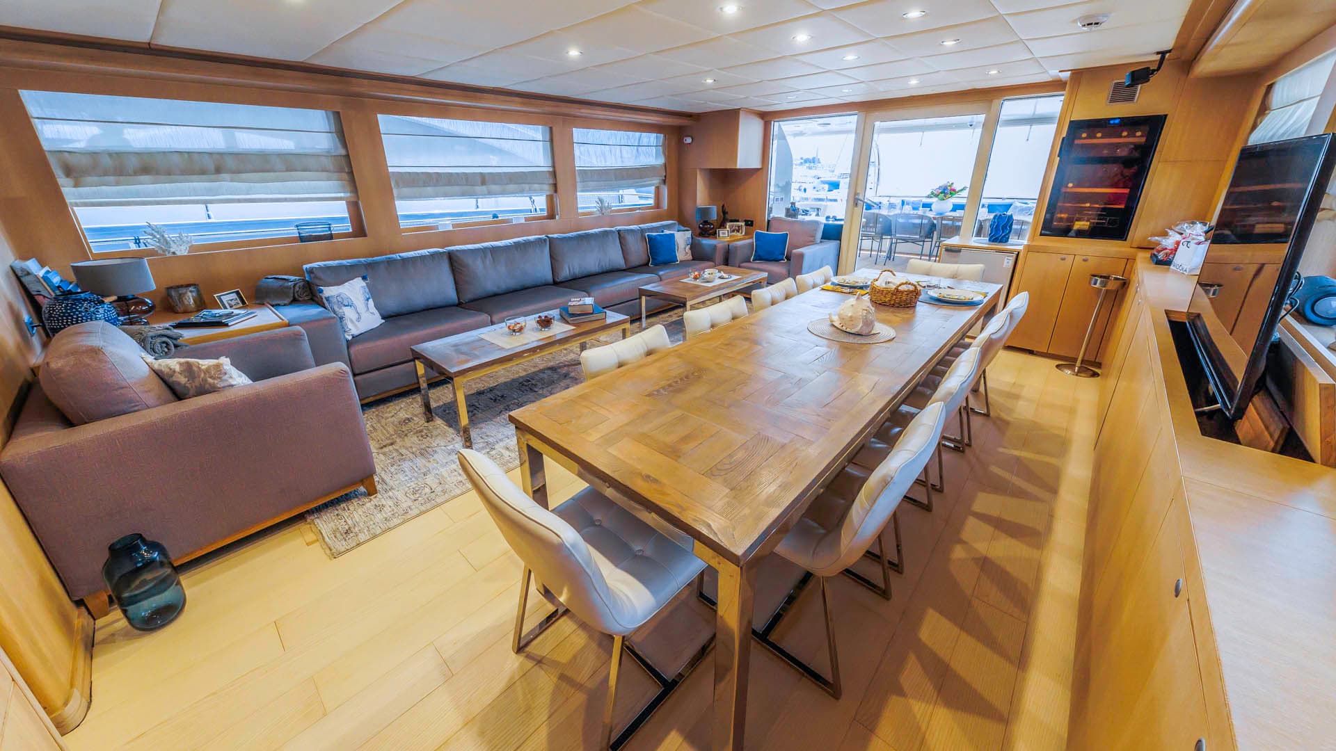 Yacht interior with wooden dining table, white chairs, and lounge area overlooking the water.