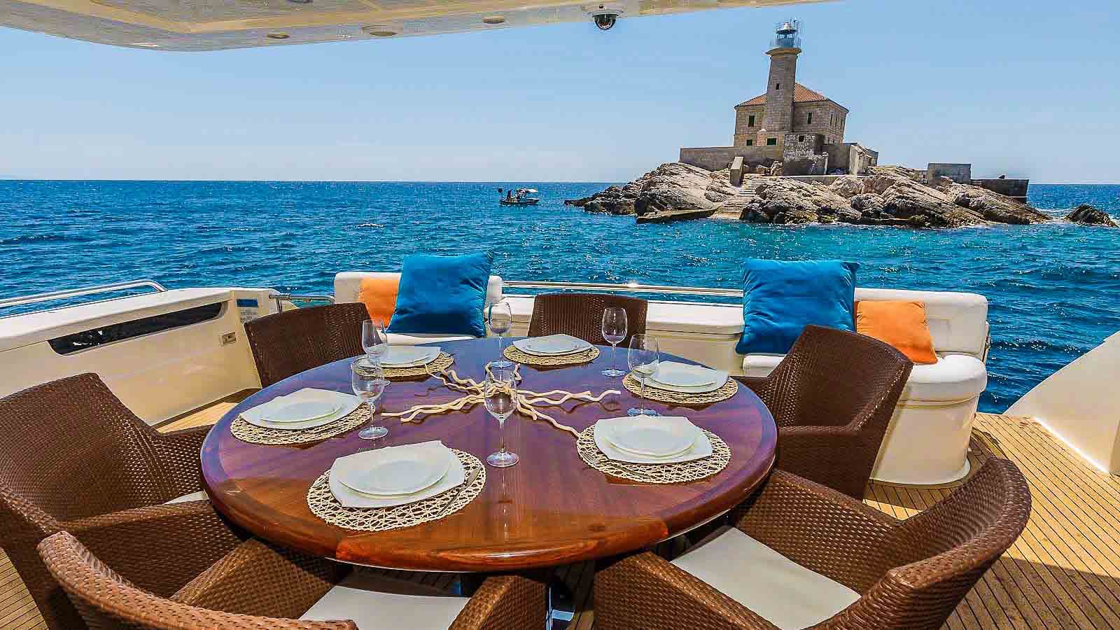 Outdoor dining setup on yacht deck overlooking rocky island with lighthouse in blue sea