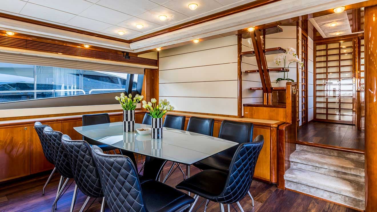 Dining area inside a yacht with wood paneling, glass table, and black quilted chairs.