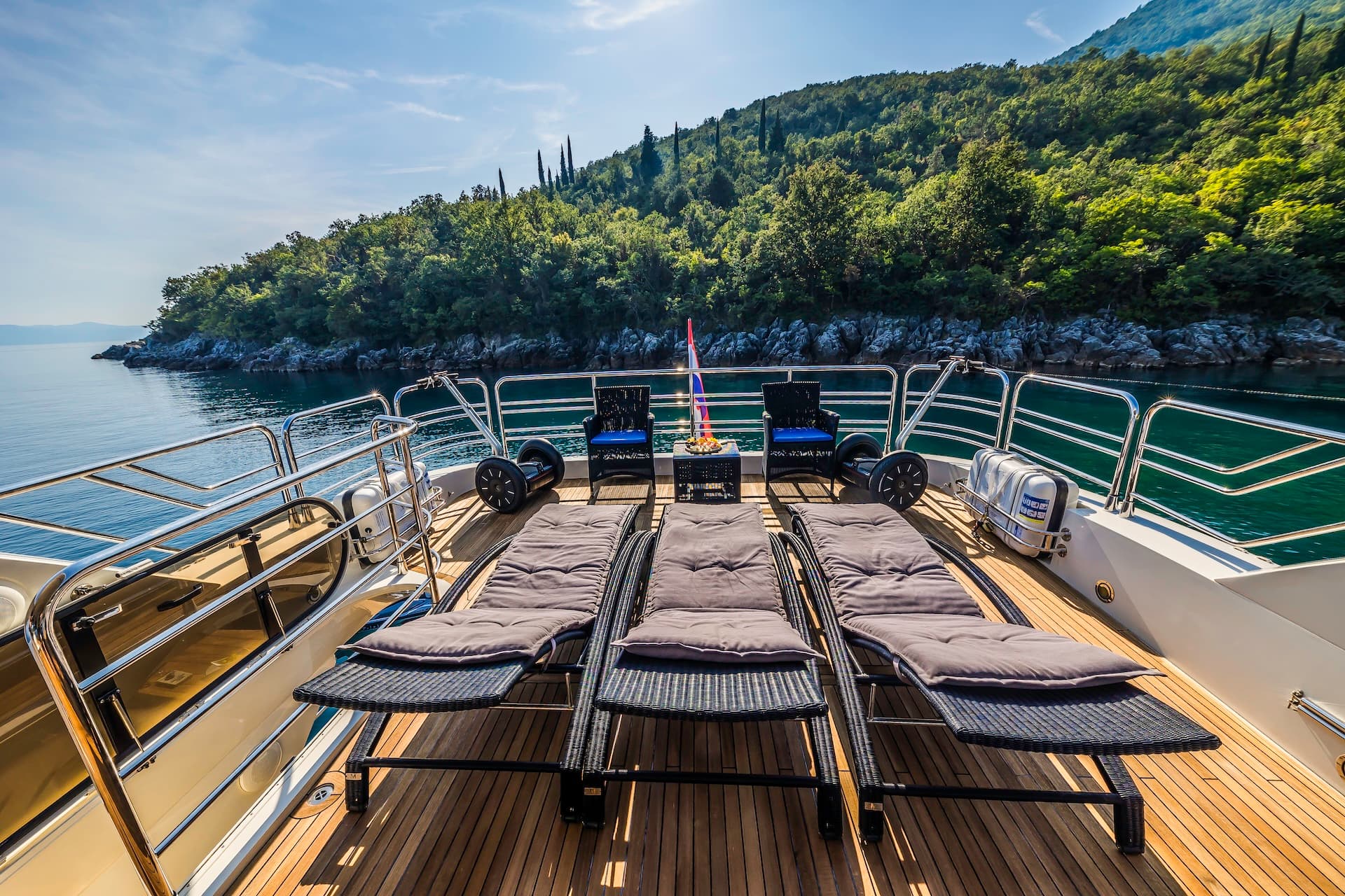 Yacht deck with lounge chairs near tree-covered coastline on sunny day