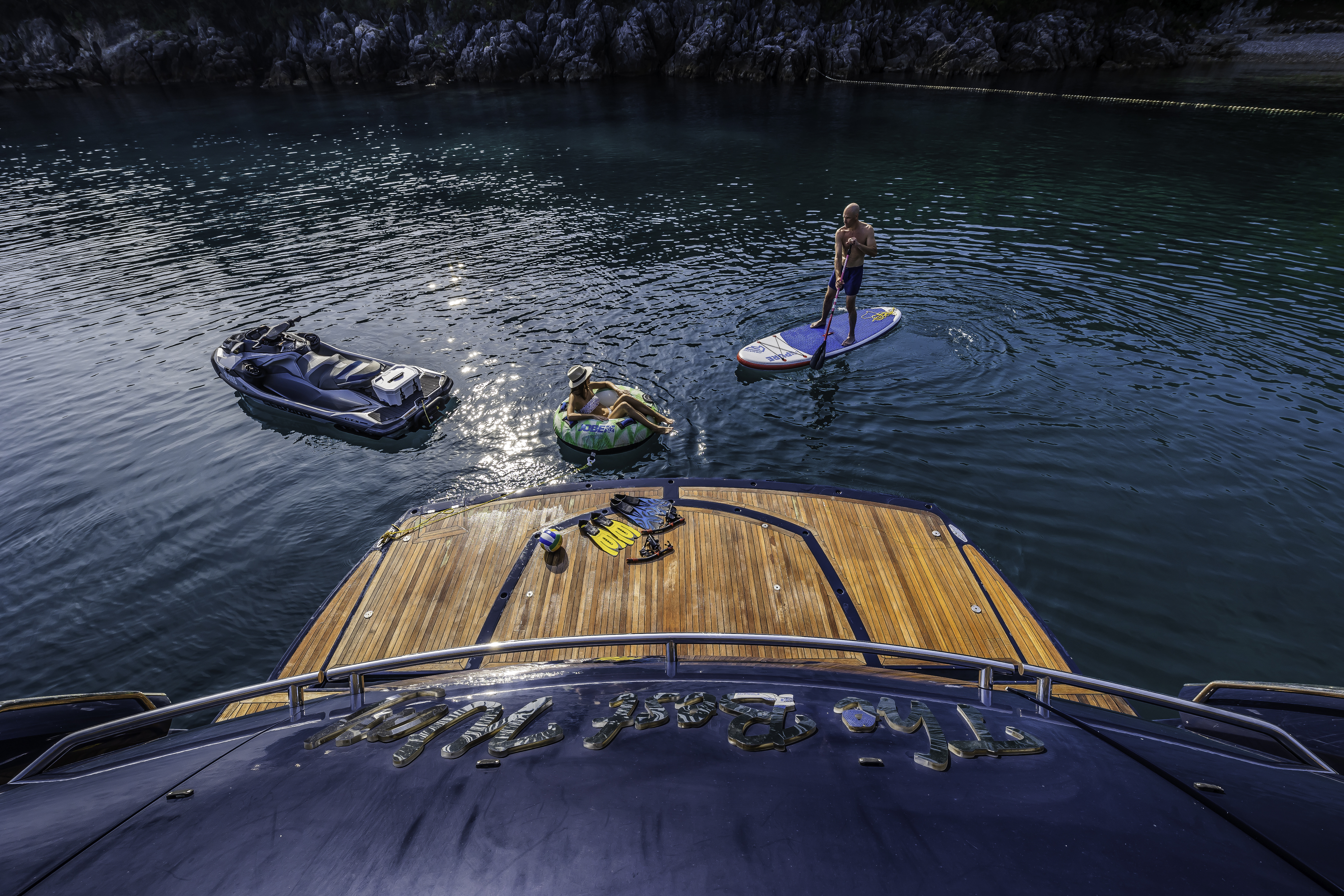 People enjoying water sports near a yacht with teak deck, rocky coastline in background
