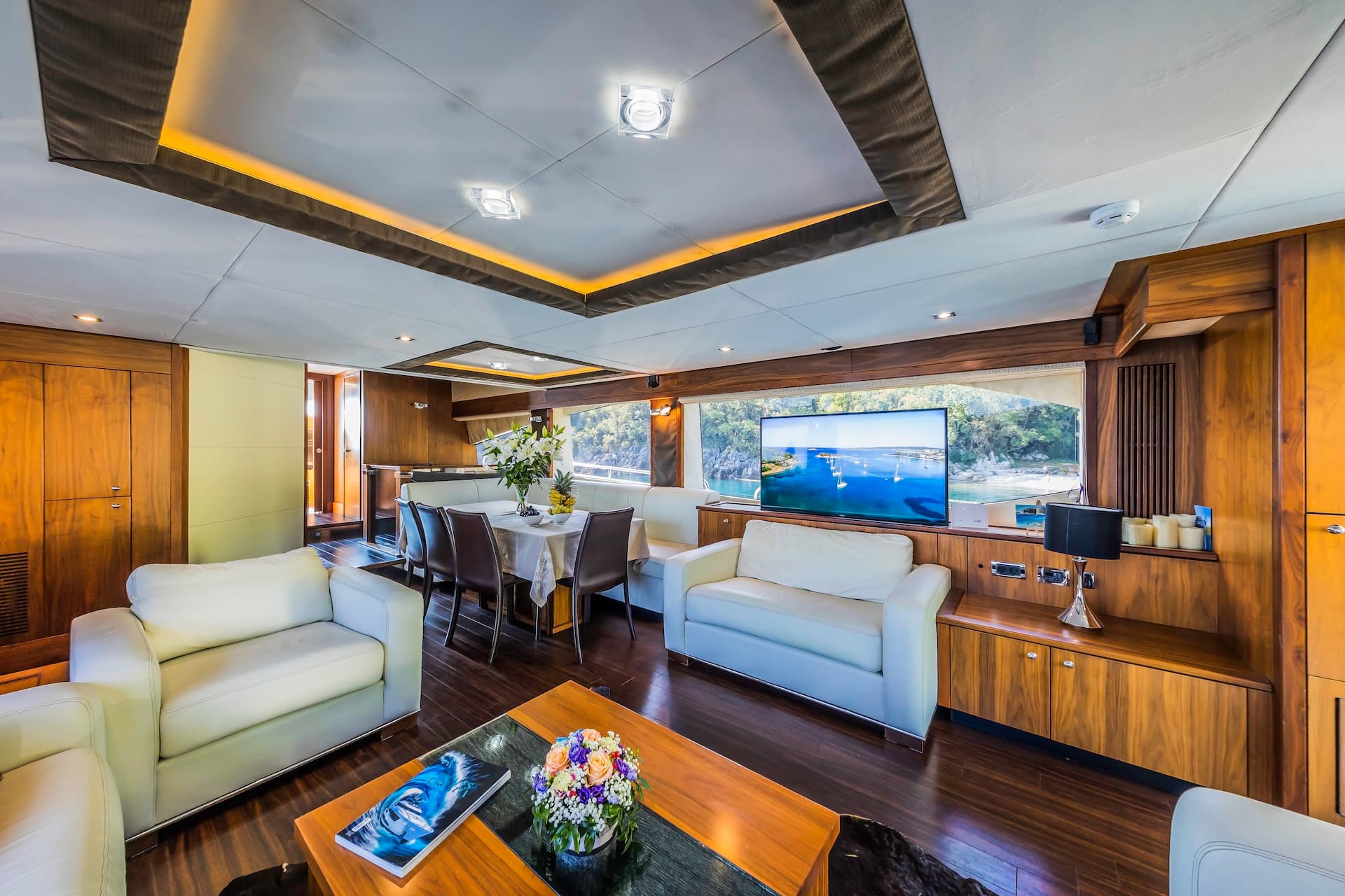 Luxury yacht saloon with white leather seating, wood paneling, and view of forested coastline.