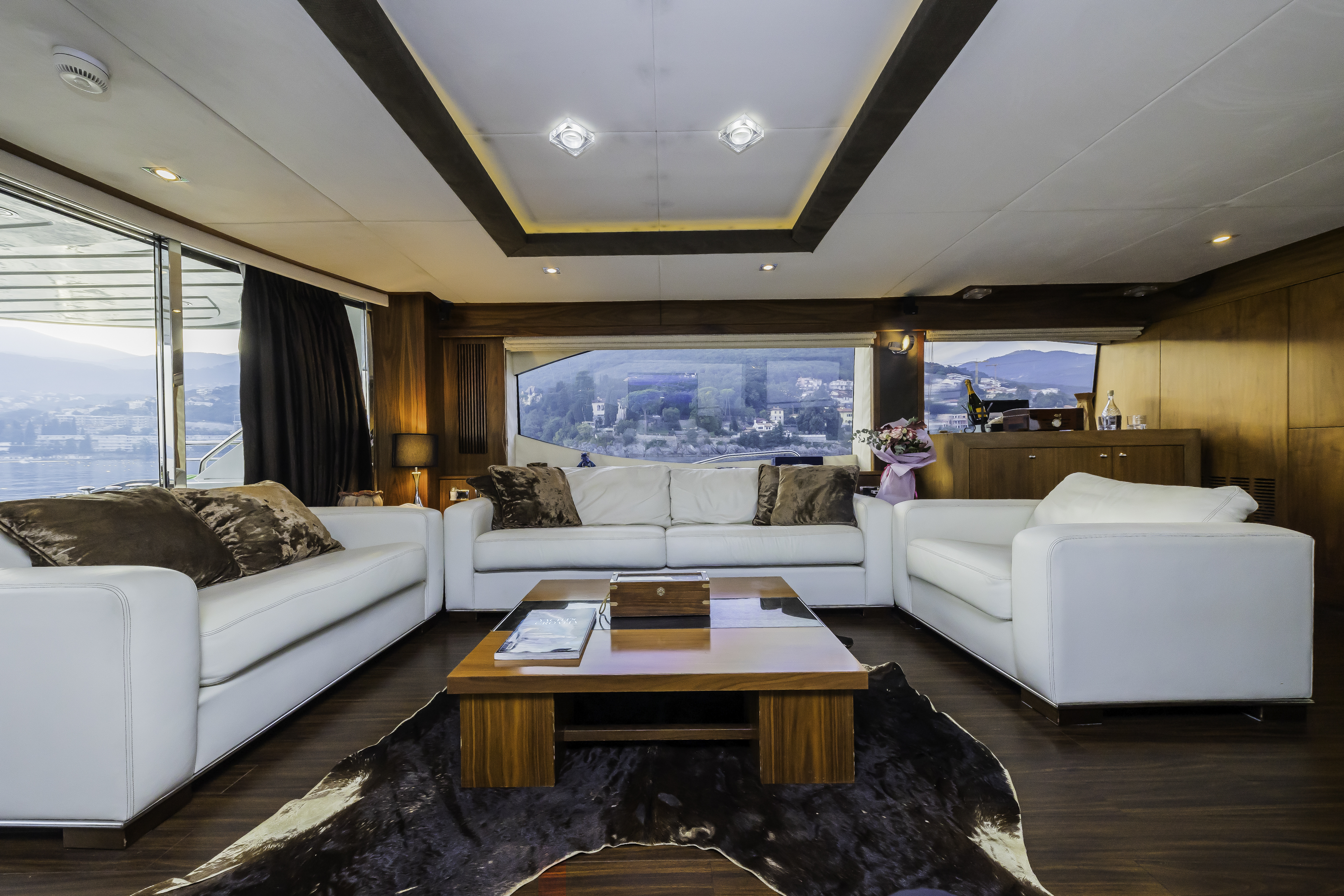 Luxury yacht interior with white sofas, wood paneling, and view of coastal hills.