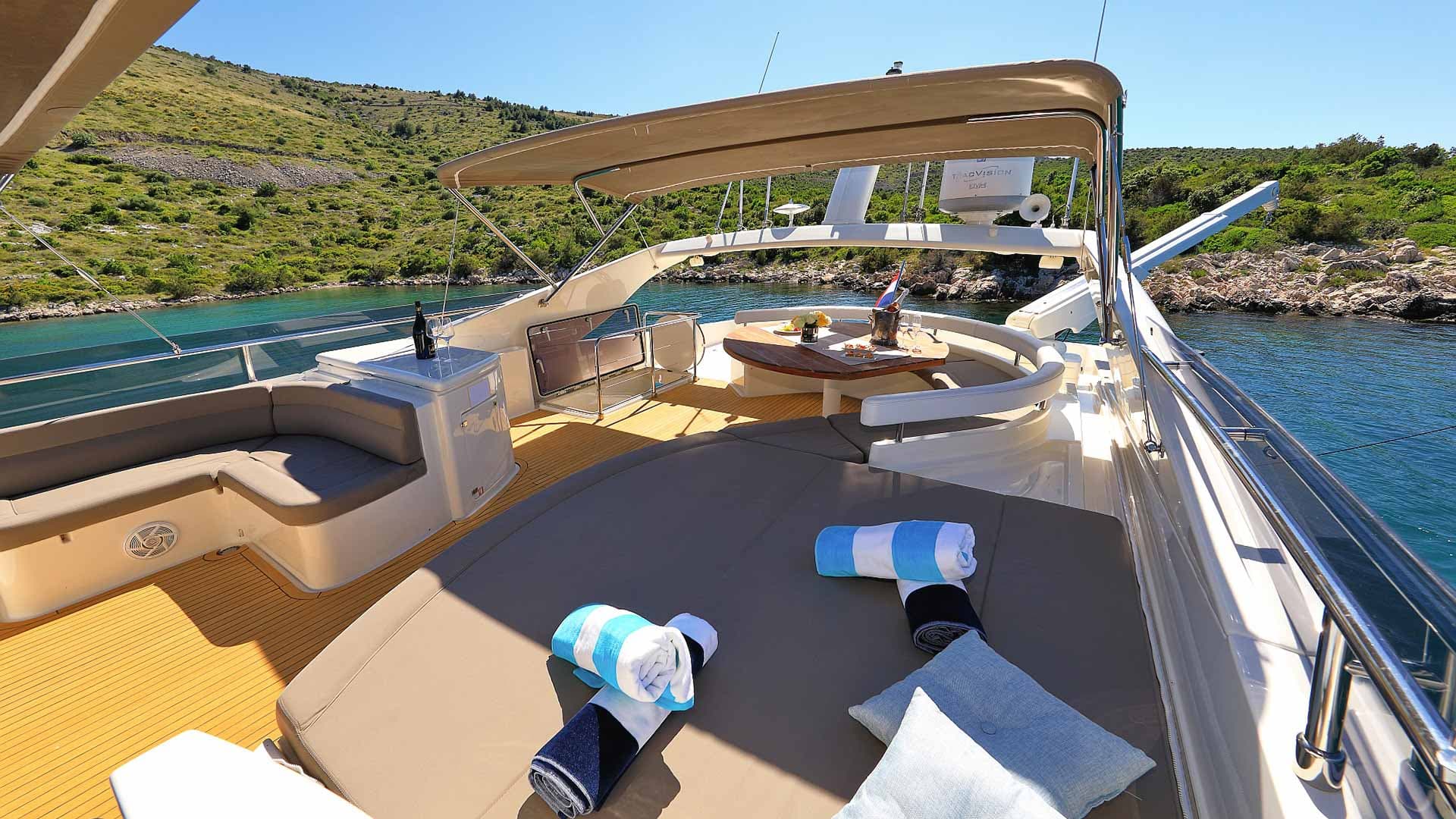 Yacht deck with sun pads, dining table, and towels anchored near a lush, rocky coastline.