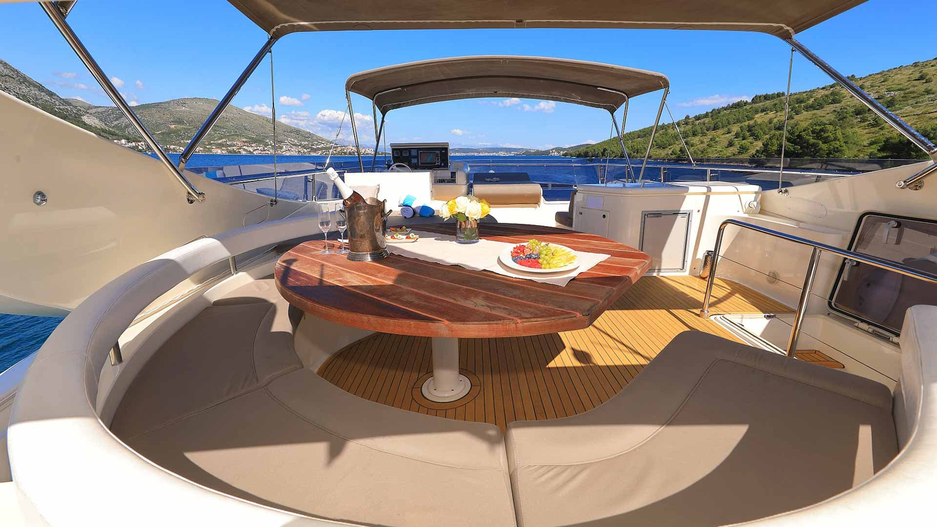 Yacht dining area with fruit and champagne overlooking green hills and blue sea.