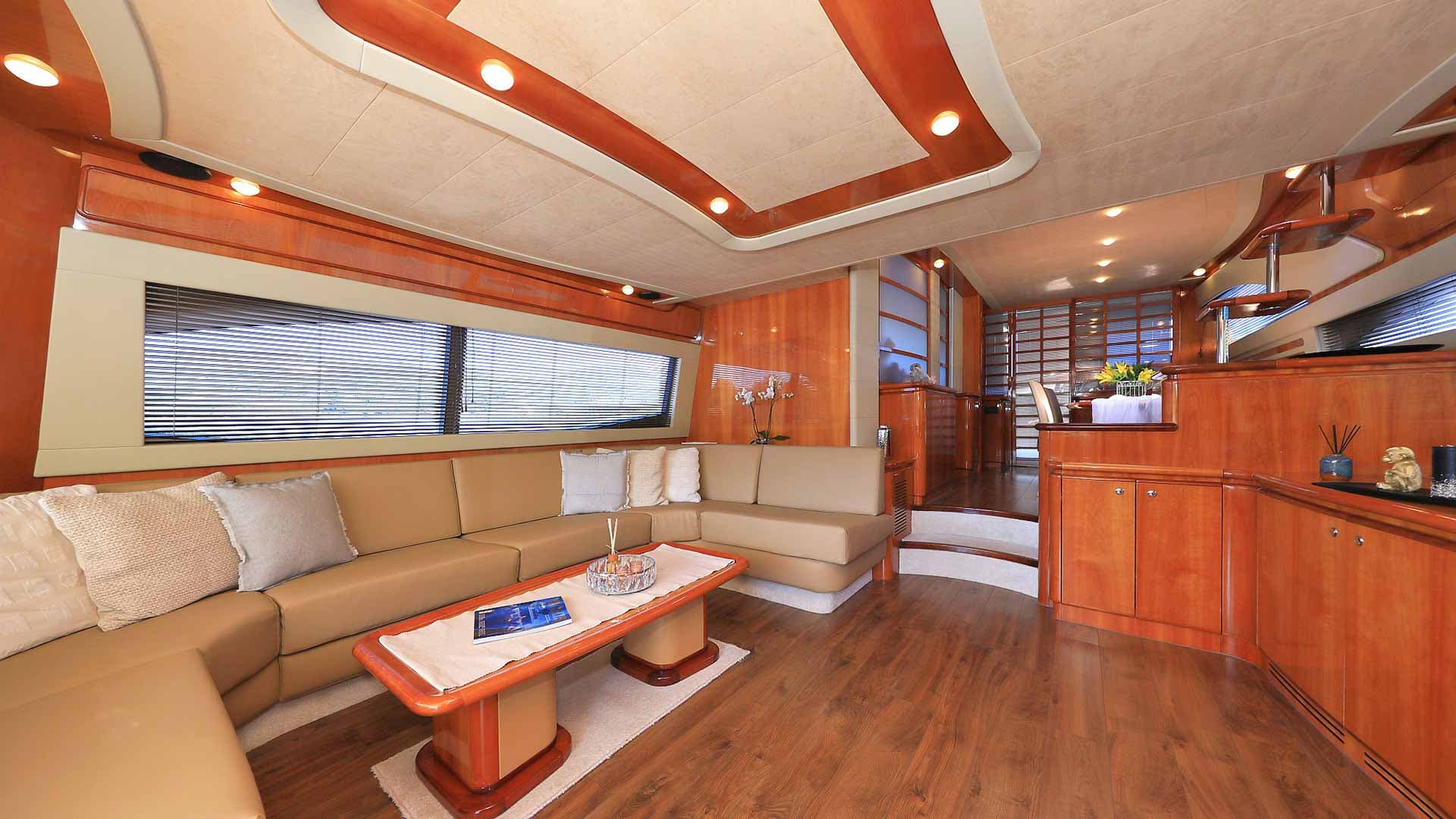 Interior salon of a yacht with tan sectional sofa, wood paneling, and bright overhead lighting.