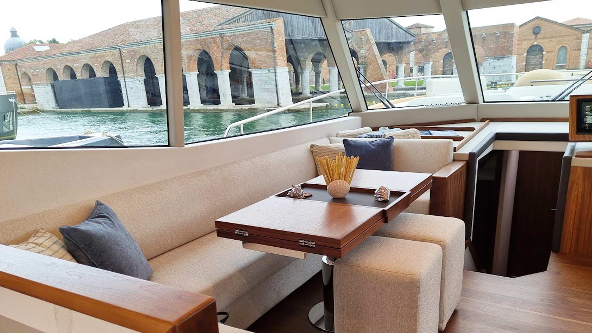 Yacht interior lounge with wooden table, looking out at historic brick buildings in water.
