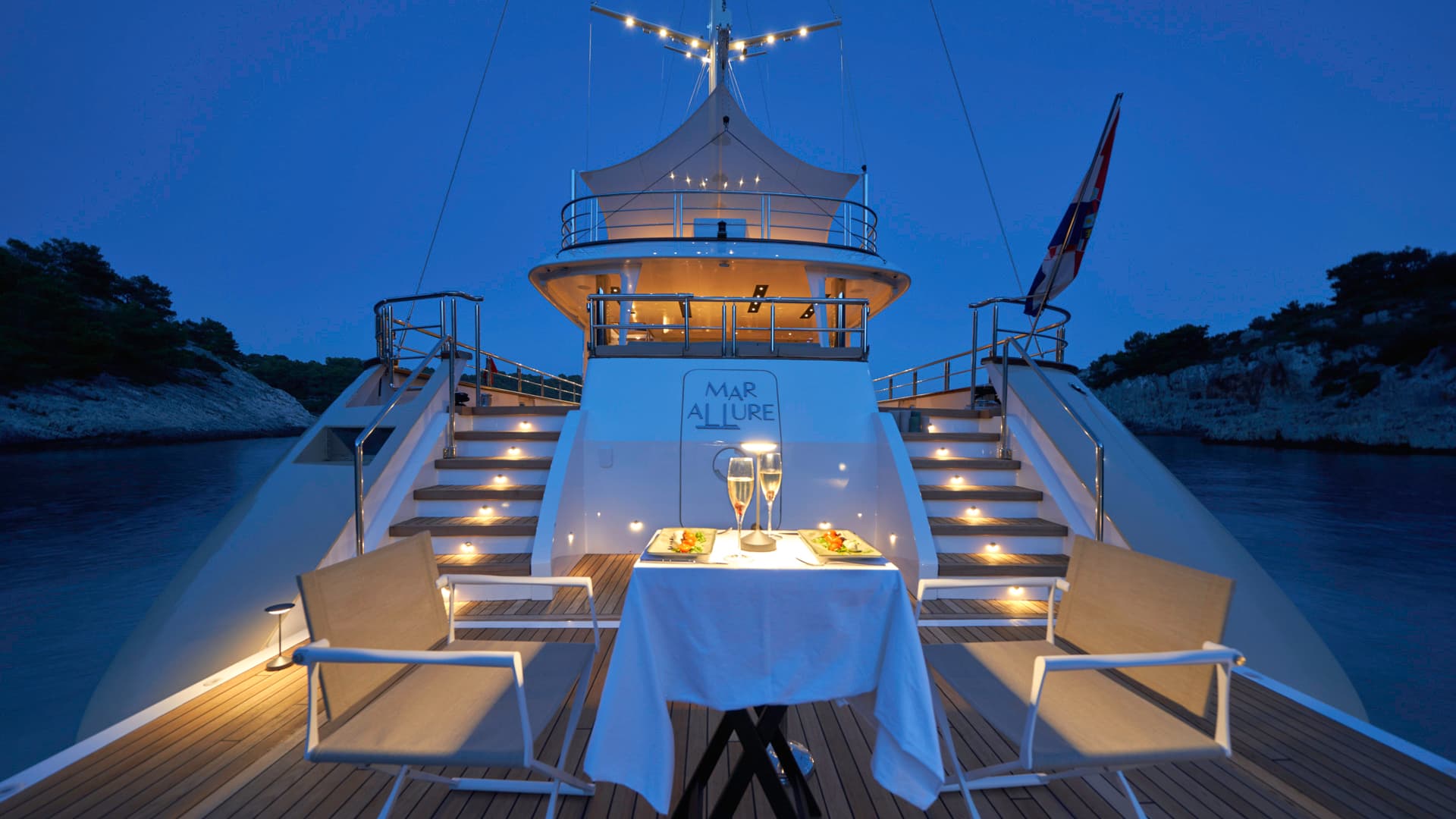 Yacht stern deck set for romantic dinner with champagne at night near wooded cliffs.