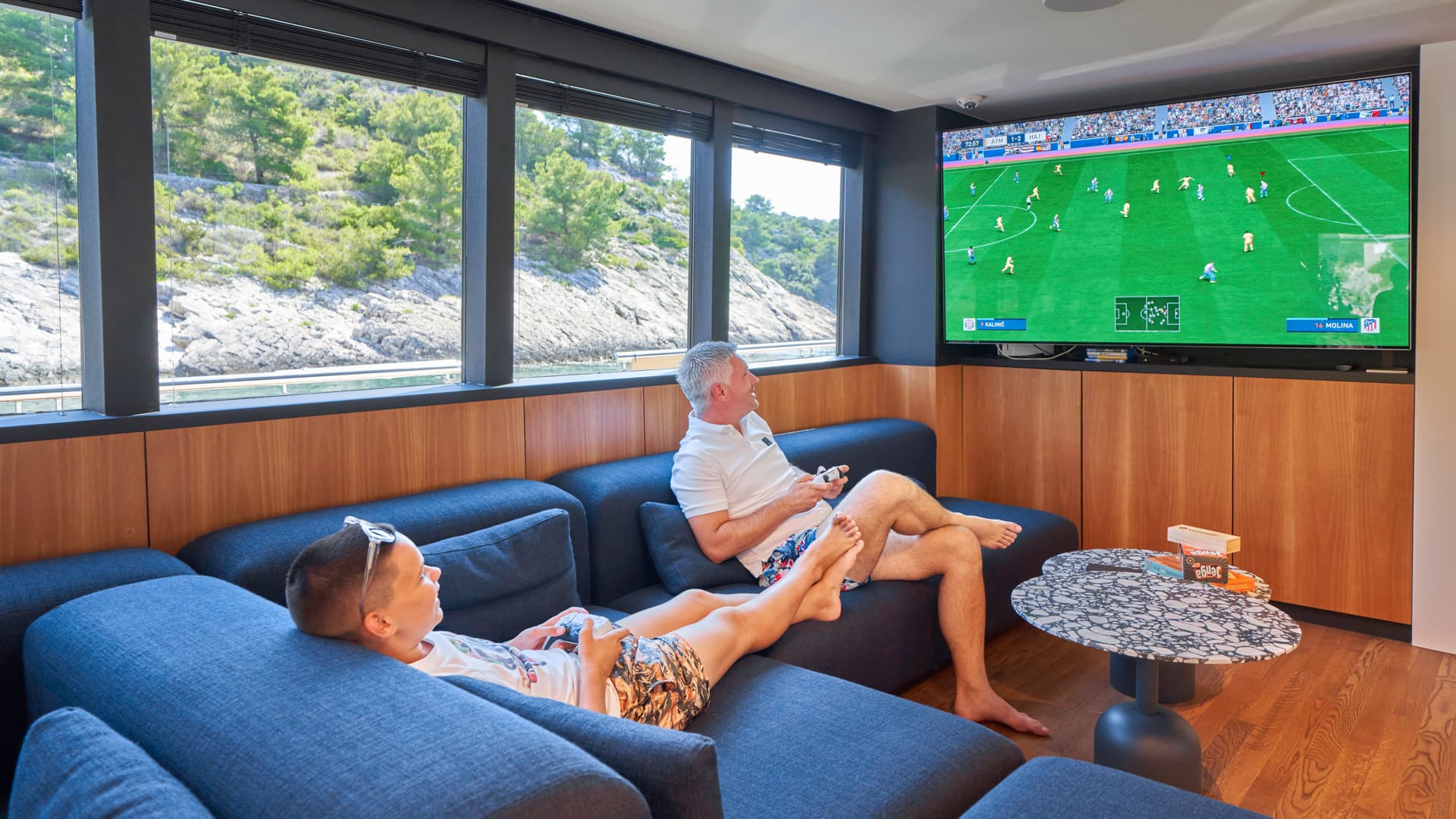 Man and boy playing video games inside yacht lounge with view of rocky, green coastline.