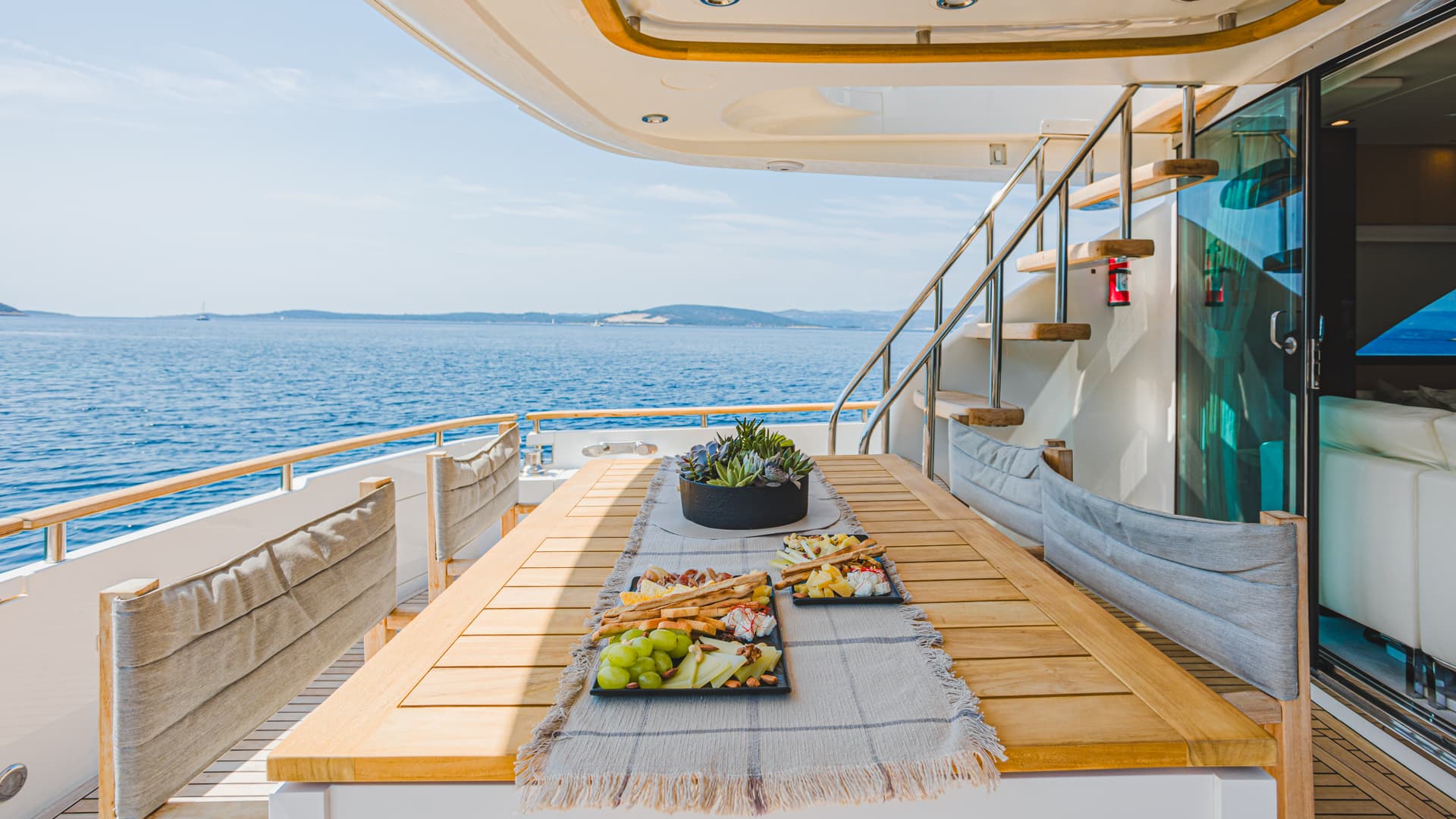Outdoor dining table with cheese and grapes on a yacht deck overlooking blue sea and distant islands.