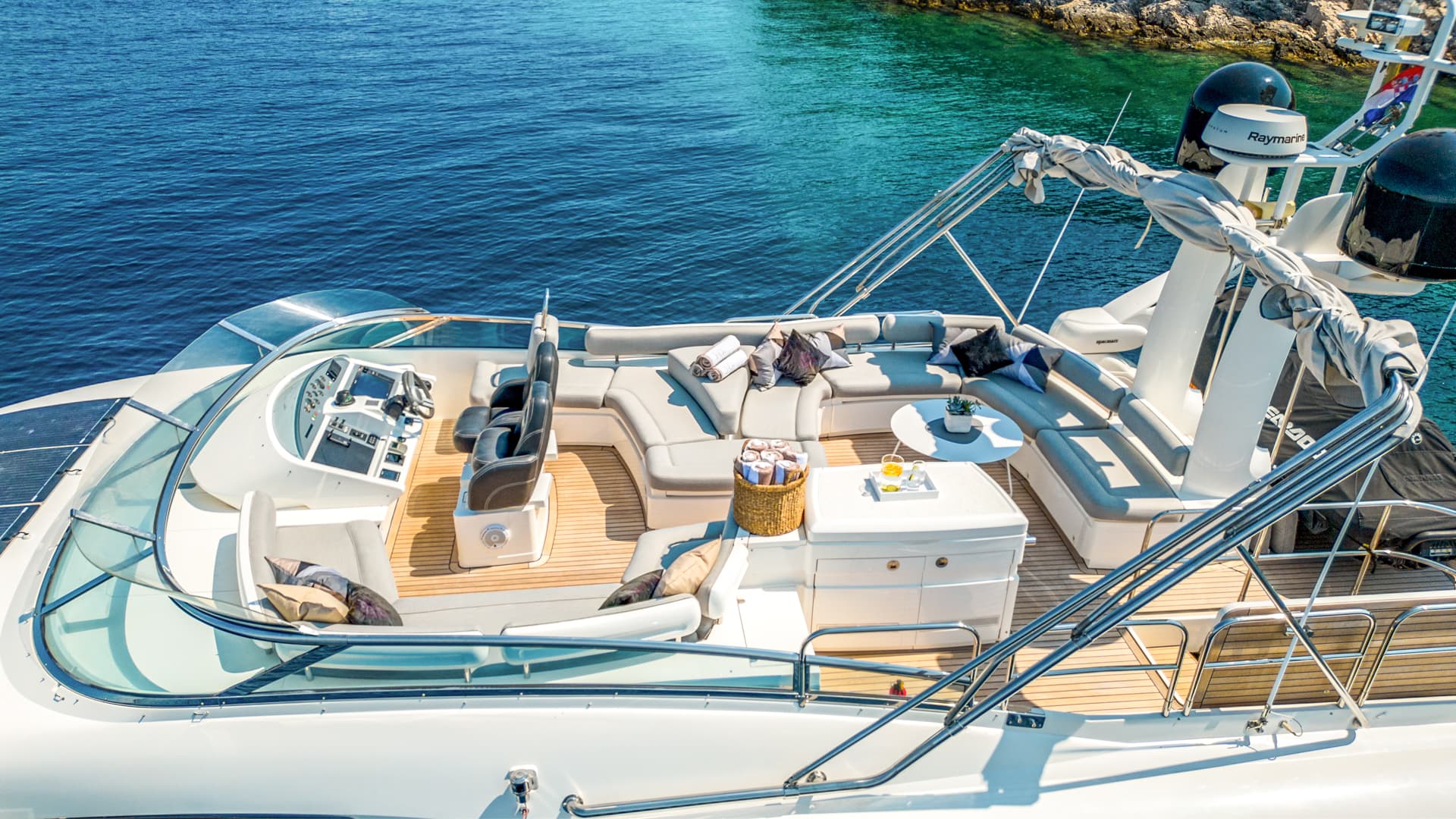 Flybridge seating area of a white yacht with teak decking near rocky coastline.