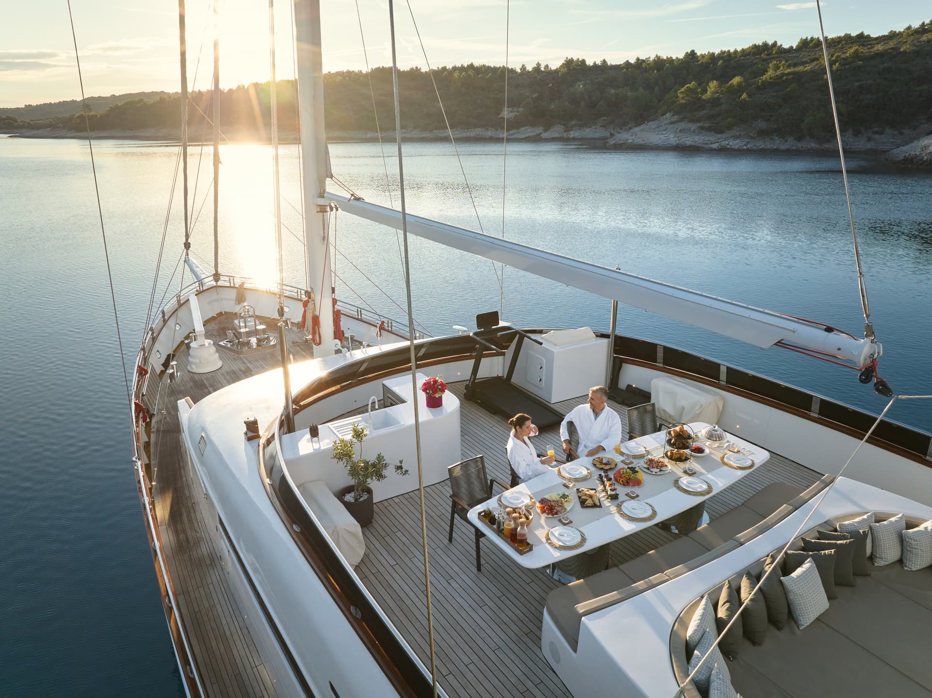 Couple in robes dining on yacht deck near forested coastline at sunset