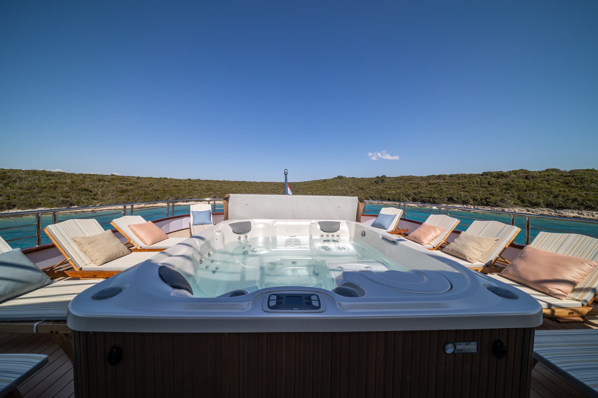 Jacuzzi on yacht deck with sun loungers, overlooking green coastline and turquoise water.
