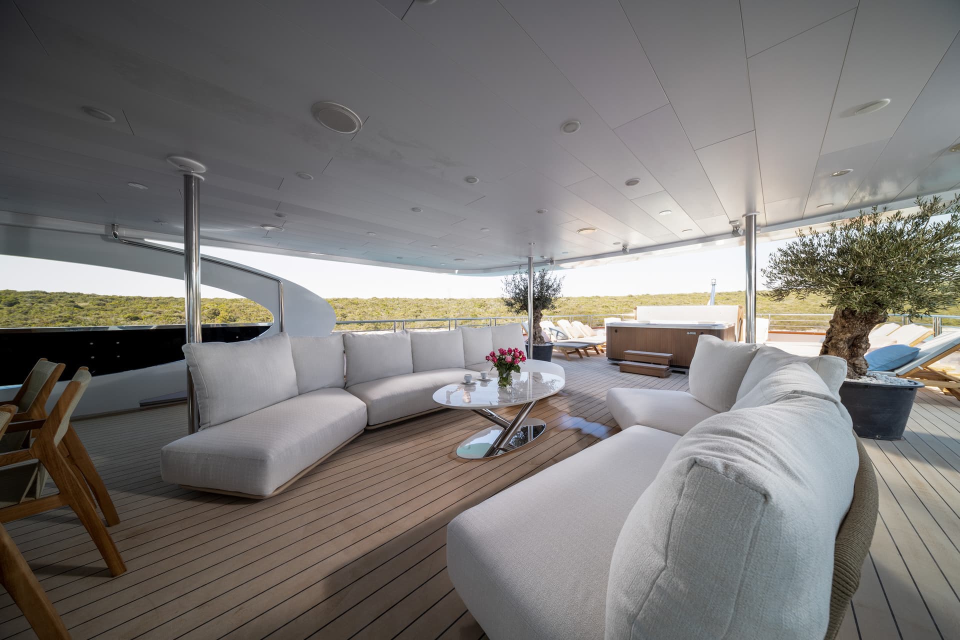 Outdoor lounge area on a yacht deck with white sofas, coffee table, and hot tub overlooking green coastline.