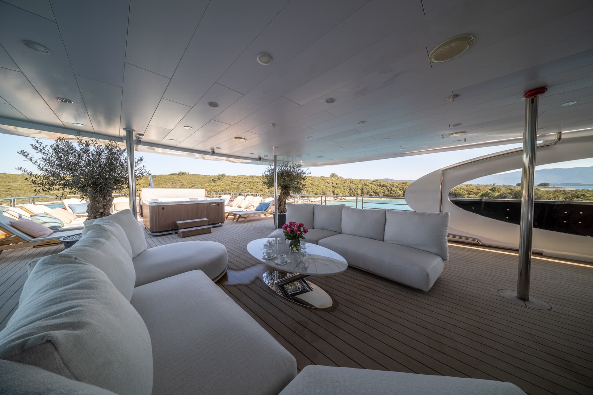 Luxury yacht deck lounge with hot tub, white sofas, and view of tree-covered coastline.