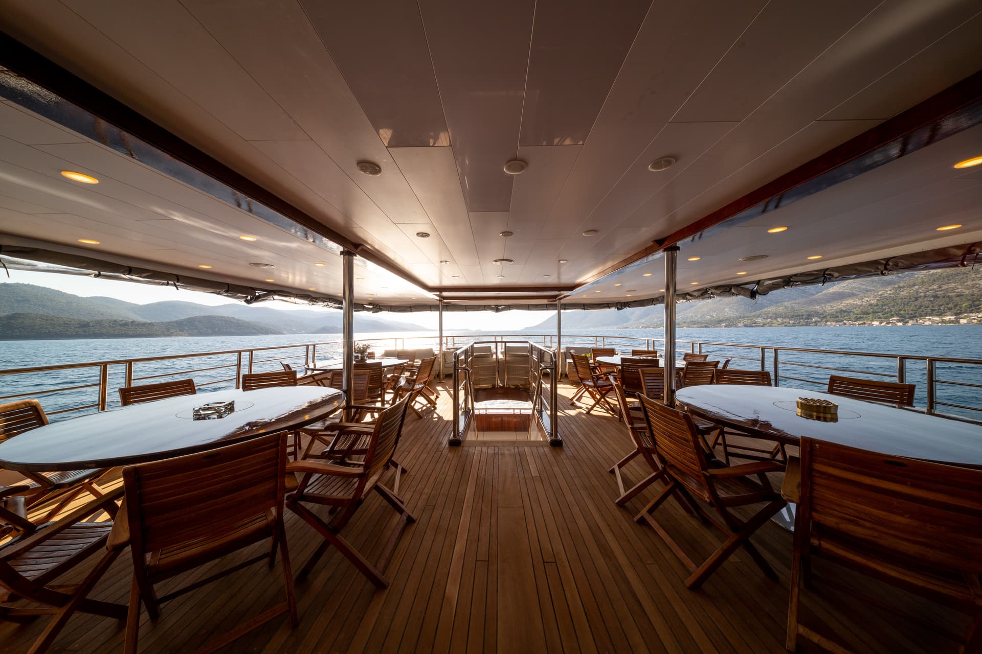 Outdoor seating area with wooden tables and chairs on a boat deck overlooking water and hills.