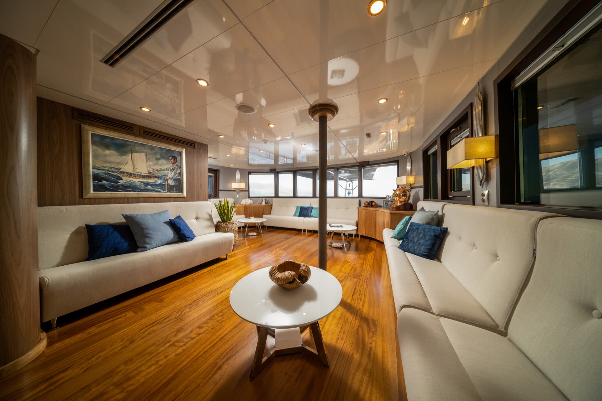 Lounge interior of a yacht with white couches, wood floors, and a painting of a sailboat.
