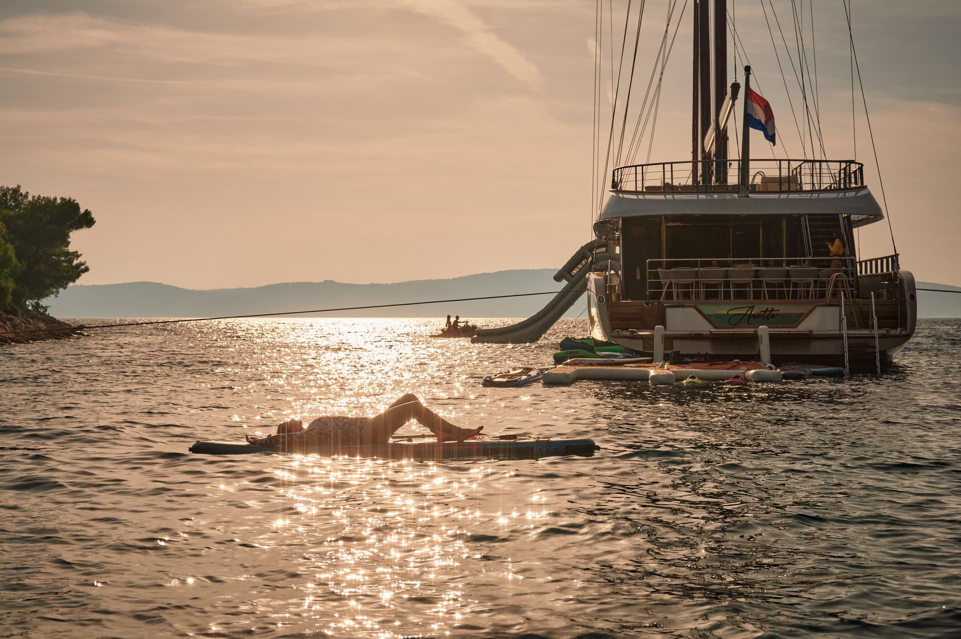 Person sunbathing on paddleboard near large yacht at sunset on sparkling water